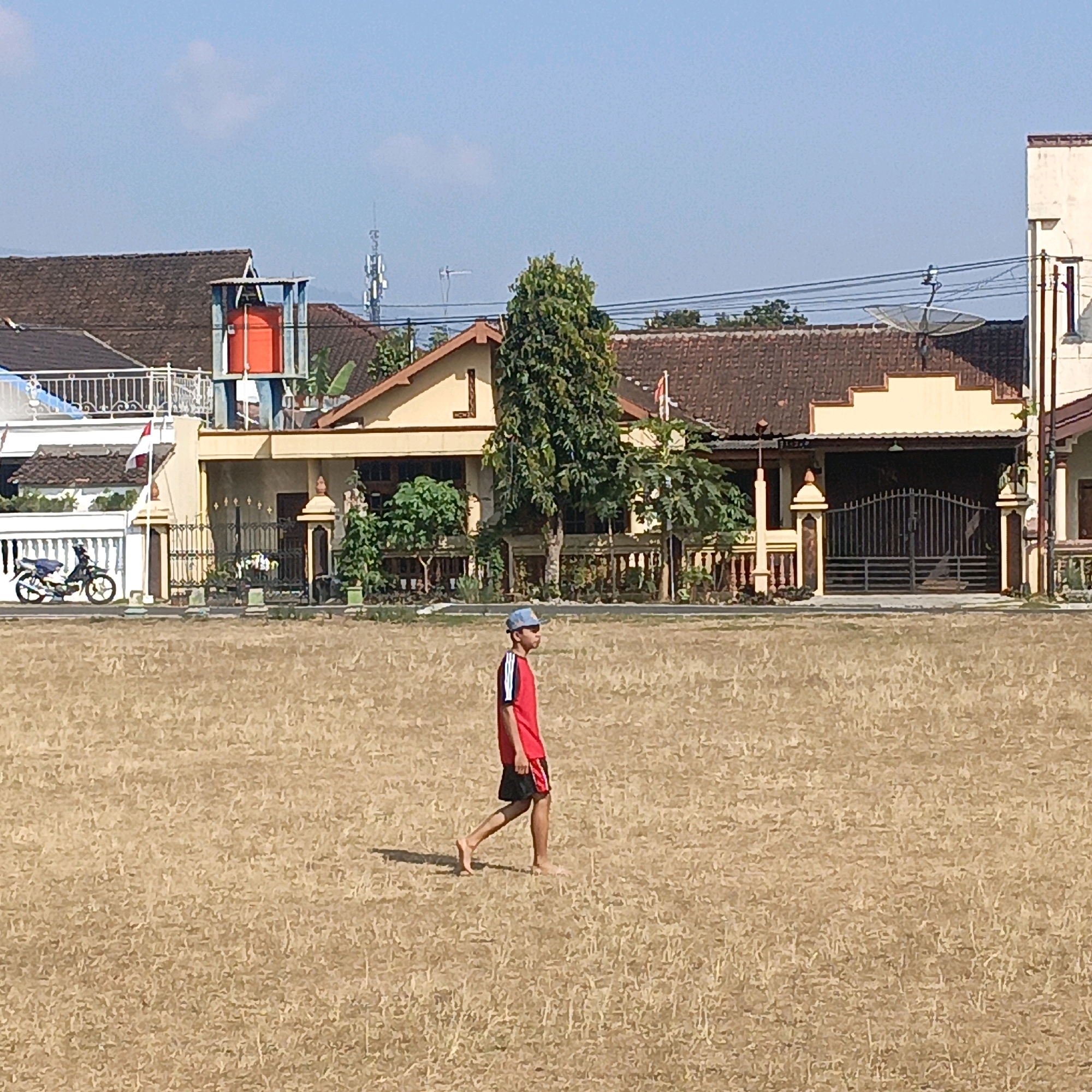 The image depicts a person walking across a dry, grassy field under a clear blue sky. The person is a male, wearing a red short-sleeved shirt with white and black stripes on the sleeves, and black shorts with red stripes. He is wearing a blue baseball cap and appears to be barefoot. The field is large and appears to be an open space, possibly a sports field or park, with dry, yellowed grass.
In the background, there are several buildings with tiled roofs, suggesting a residential area. Some of these buildings have fences and gates, and one building has a water tank on its roof. A motorcycle is parked near a white fence in the left background. There are also several trees scattered throughout the scene, and overhead wires are visible. The overall impression is of a sunny day in a semi-urban or suburban environment in Salatiga, Indonesia.
