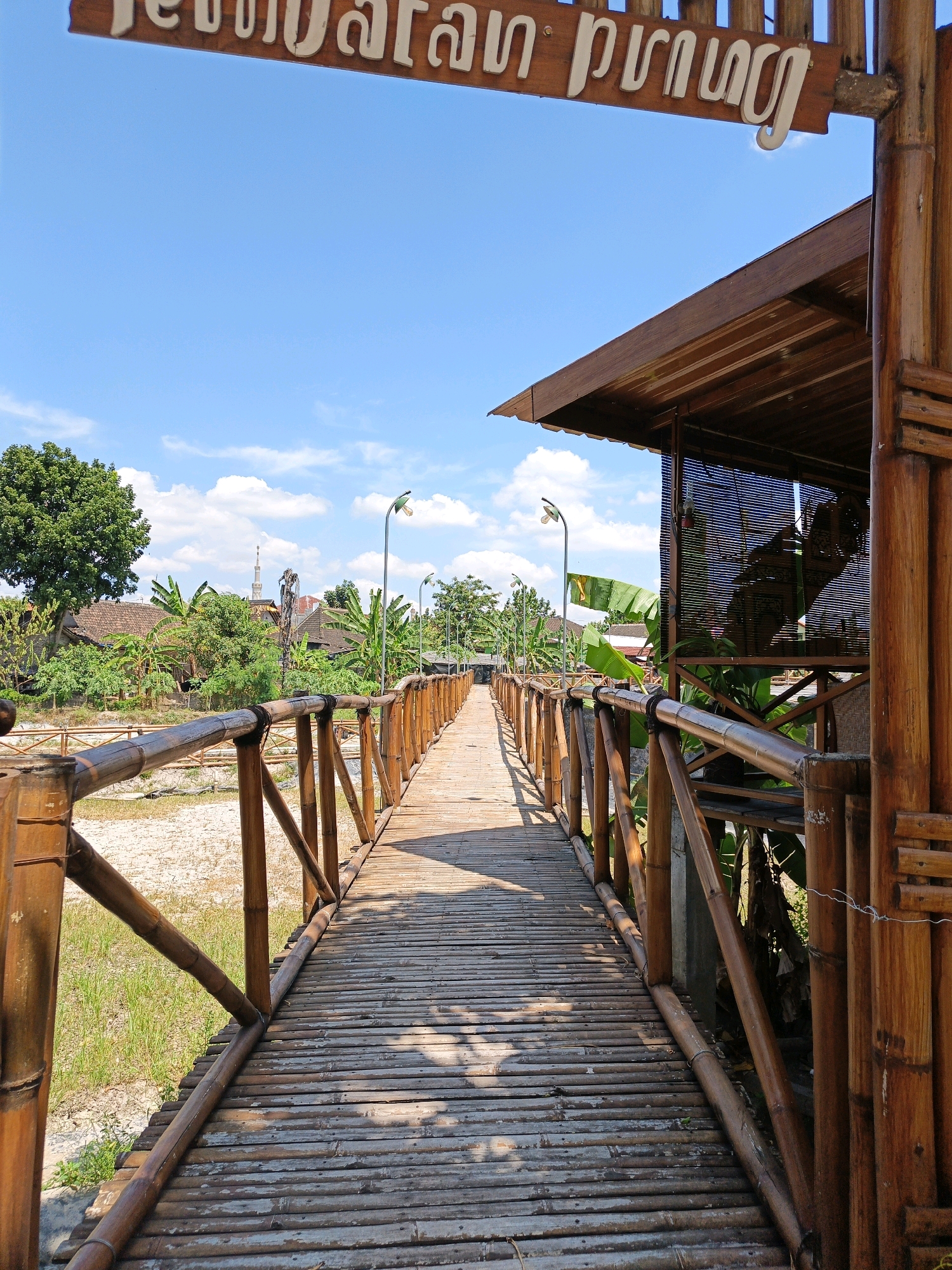 The image shows a bamboo bridge that leads to a building with a sloped roof, made of the same material. The bridge is lined with bamboo railings. The environment appears to be rural, with trees and houses visible in the background. The sky is blue with some clouds, and the sun is shining, casting shadows on the bridge. A sign is visible at the top of the image, with text that appears to be in Indonesian. The location is identified as Jonggrangan, Indonesia. There are no people or animals visible in the image. The bridge itself is the main subject, along with the surrounding natural and architectural elements.