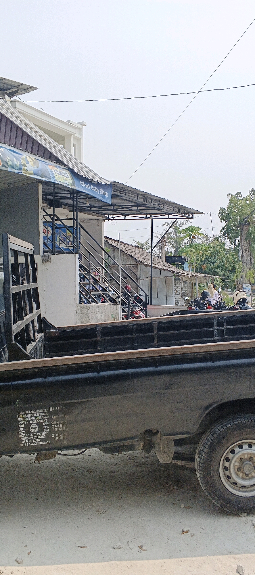 In Sonoadi, Indonesia, a pickup truck is parked on a dusty road in front of a shop. The shop, identified as "Alfart Baby Shop" by a banner, has a metal awning extending over its entrance, which features a set of stairs leading up to it. Several motorcycles are parked in front of the shop and on the stairs. A woman wearing a hijab is visible near the motorcycles. The pickup truck's side displays text and numbers, including "GVW 2540KG", "CPW 2420KG", and "PASSENGERS 3 ORANG". The environment is outdoors with clear skies and trees in the background.