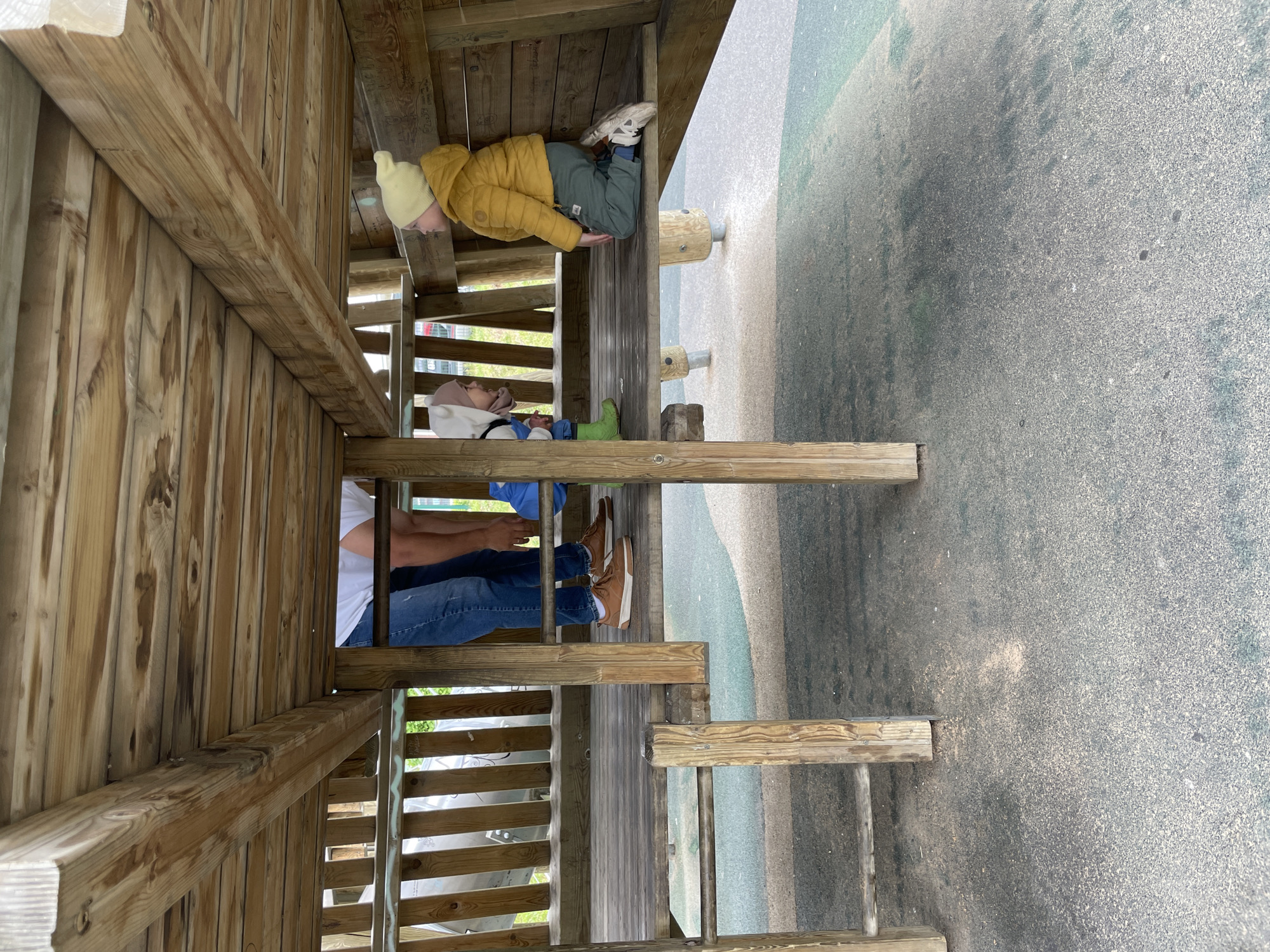 The image depicts an outdoor scene within a multi-level wooden playground structure. Three individuals are present across different levels of the play area.
On an upper level, two children are visible. One child is dressed in a yellow puffer jacket, a light yellow knitted beanie, and grey-green trousers with white sneakers. This child is seated on a sturdy horizontal wooden beam, facing away from the viewer and appearing to look towards the side or down. Behind them, further back into the structure, another child is seated on the wooden platform. This child wears a white hooded top with ear-like details, light blue trousers, and distinct green boots.
Below the children, on a lower section of the wooden structure, an adult is partially visible. The adult wears a white t-shirt, blue jeans, and brown lace-up shoes, with only their torso and legs visible. They appear to be seated or standing on the lower platform, supervising or resting.
The playground itself is constructed from natural wooden planks, posts, and beams, which show signs of use and weathering. Faint, unreadable scribbled markings are visible on some of the wooden surfaces. The ground beneath and surrounding the playground is a grey and greenish-speckled safety surface, likely made of rubber mulch or similar soft material, designed to cushion falls.
The scene takes place during daylight hours, with soft, diffused lighting suggesting either an overcast sky or late afternoon/early morning conditions. The activity appears to be casual recreation and play within the playground environment. No discernible text is clearly visible on any object. The location context provided is Izhevsk, Russia.