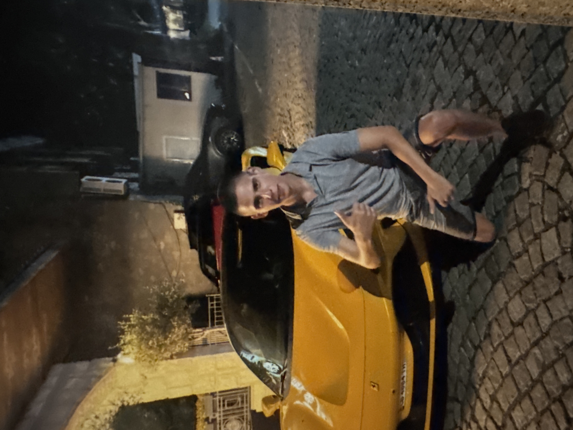 The image depicts a young man posing next to a bright yellow sports car on a cobblestone street at night in Sofia, Bulgaria. The man, with short hair, is wearing a grey polo shirt and dark shorts. He is leaning against the yellow car, which appears to be a Ferrari, with one arm resting on its roofline and the other hand giving a thumbs-up gesture. He looks directly at the camera with a neutral to slightly smiling expression, suggesting a casual and relaxed mood.
The setting is an urban street, characterized by the dark cobblestone paving and background buildings. Behind the yellow sports car, parts of a dark-colored sedan or hatchback are visible. Further back, several buildings can be seen, some with visible windows, indicating an older architectural style. An outdoor utility box or air conditioning unit is present on a wall. A small, bushy plant or tree is also discernible near a building wall. The scene is illuminated by artificial light sources, casting shadows and confirming the nighttime setting. The ground appears dry. Faint, unreadable text is visible on a small sticker or label on the side of the yellow car.