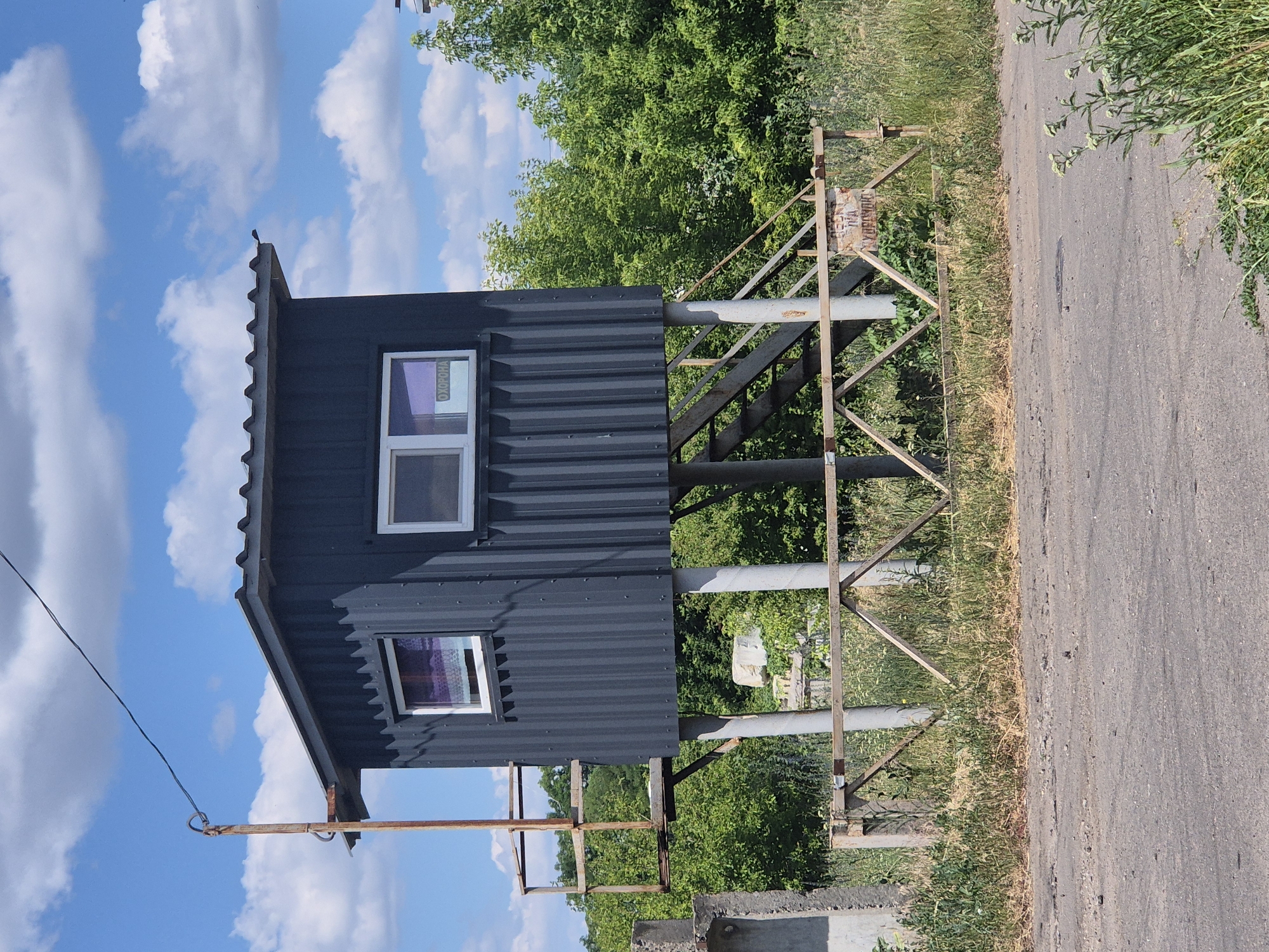 A dark gray, corrugated metal guard booth stands elevated on four cylindrical concrete pillars in Sozonivka, Ukraine. The booth has a sloped roof and two visible windows with white frames. Through one window, a purple curtain is visible. The other window has a sign that reads "ОХОРОНА" (Ohorona), meaning "Guard" or "Security" in Ukrainian. A metal staircase with railings leads up to the booth. A rusty metal structure, possibly a former guard platform or a support for an antenna, is attached to the left side of the booth. In front of the booth, a simple wooden barrier with metal supports crosses a gravel and dirt path. The area is surrounded by lush green trees and foliage. The sky above is blue with scattered white clouds. The overall scene appears to be in a rural or semi-rural setting.