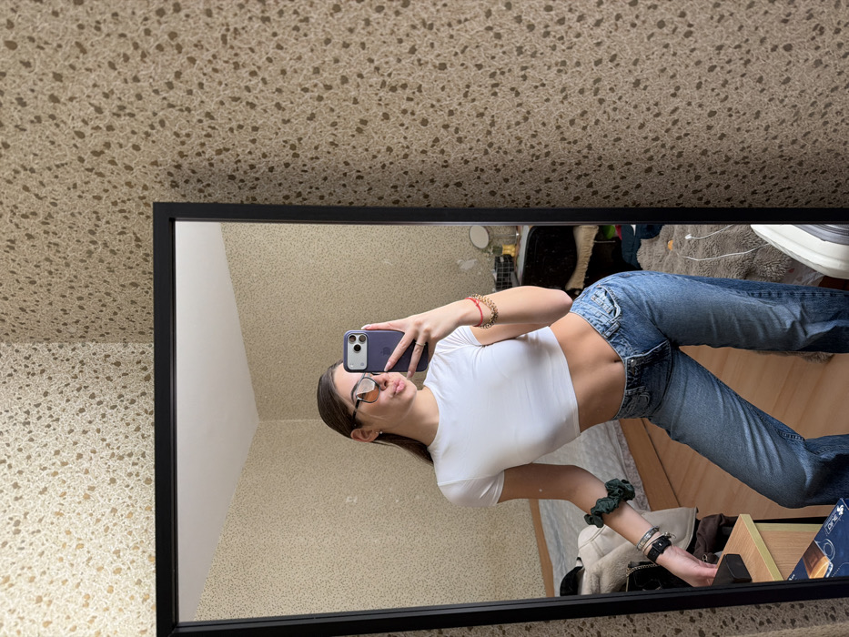 The image depicts a mirror selfie of a young female taken indoors. The subject, appearing to be in her late teens or early twenties, stands reflected in a black-framed, full-length mirror. She has long, dark brown hair and is wearing a white cropped T-shirt revealing her midriff, blue denim jeans, and dark-framed sunglasses with orange-tinted lenses. Her right arm is raised, holding a purple smartphone to capture the image. Her left wrist displays a dark green scrunchie and several bracelets, while her right wrist has a red string bracelet with gold-colored beads. Her lips are slightly parted in a pout. The reflected room features walls covered in light brown wallpaper with a textured, speckled pattern. Visible furniture includes a light-colored wooden bedside table with a white object and a blue and white box, a bed with a light-colored frame, and another dresser or vanity with various items, including a small round mirror. The floor appears to be light-colored wood or laminate. The setting is consistent with a private residential interior, such as a bedroom, and provides no specific visual indicators to determine a precise location within the city of Sofia, Bulgaria.