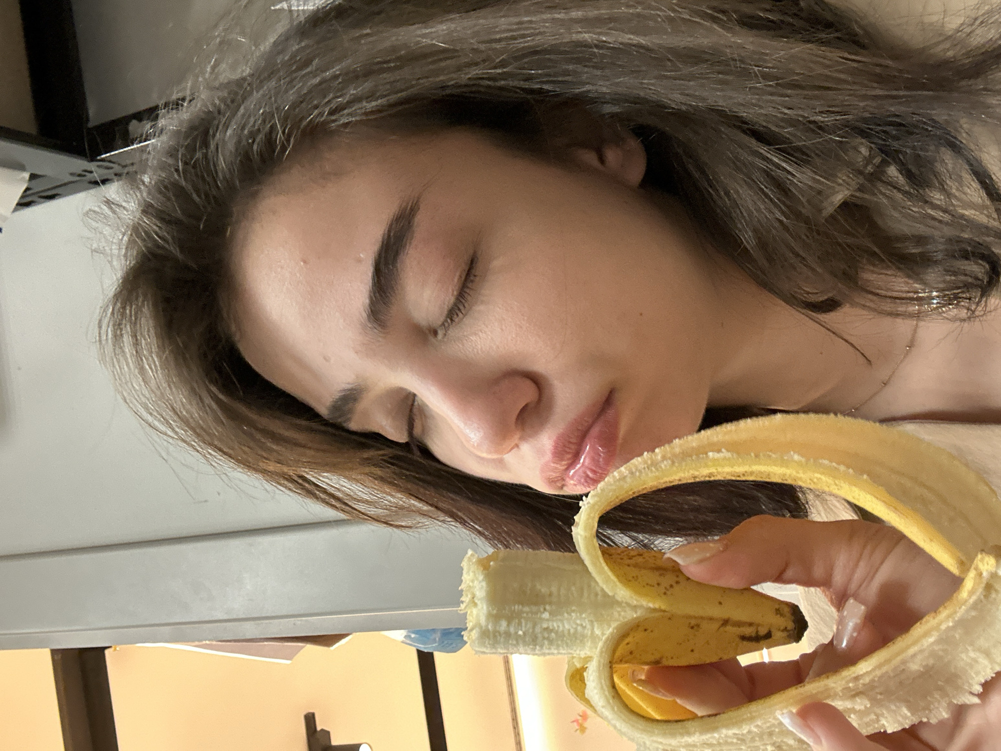 A young woman with closed eyes and light brown hair is holding a peeled banana. The banana peel is hanging open, revealing the fruit. The woman's hand has long nails. The background is blurry, showing a desk or table and a wall. The location and city of Sofia, Bulgaria cannot be confirmed from the image.