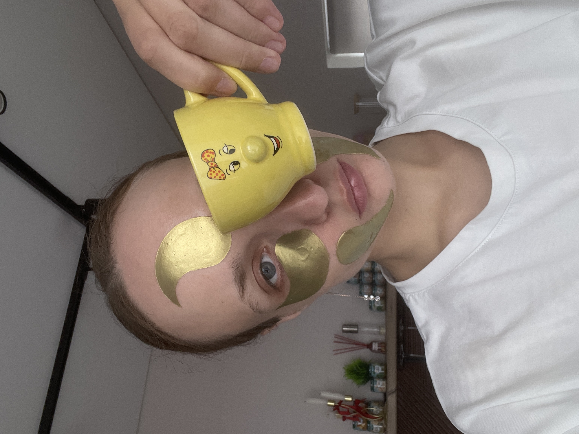 A person with gold eye masks poses indoors while holding a yellow mug. The mug has a cartoon face. The person is wearing a white shirt. A shelf is visible in the background, holding various items. The location cannot be determined definitively as Podshivalovo, Russia from the visual data alone.