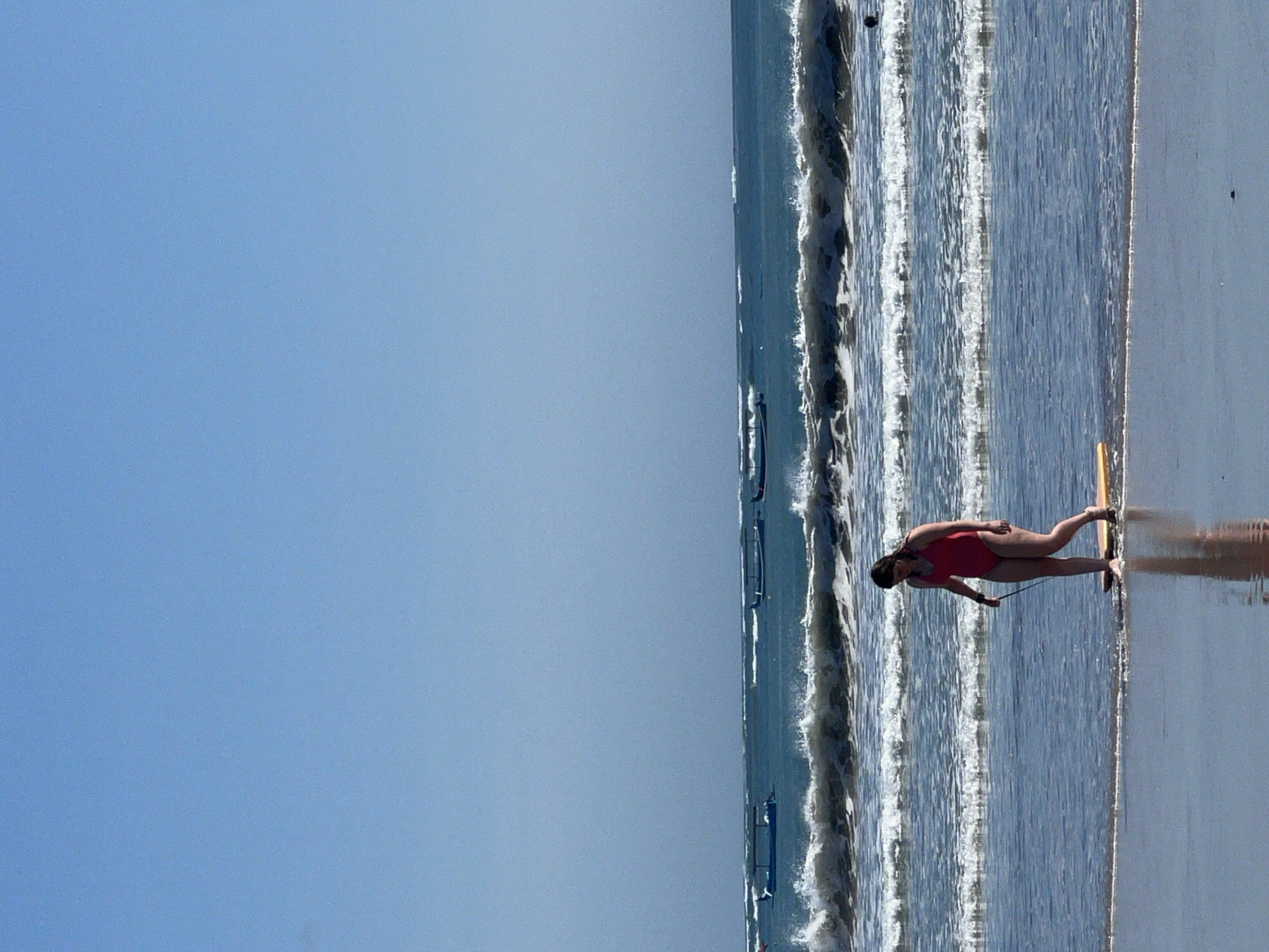 The image depicts a beach scene in Kuta, Indonesia, under a clear blue sky. The foreground features wet sand reflecting the sky and a woman wearing a red swimsuit walking in the shallow water. She is holding a leash attached to a yellow boogie board. In the middle ground, waves are breaking, creating white foam. In the background, several long, narrow boats with canopies are visible on the turquoise ocean. The overall environment is a coastal area with calm to moderately wavy conditions.