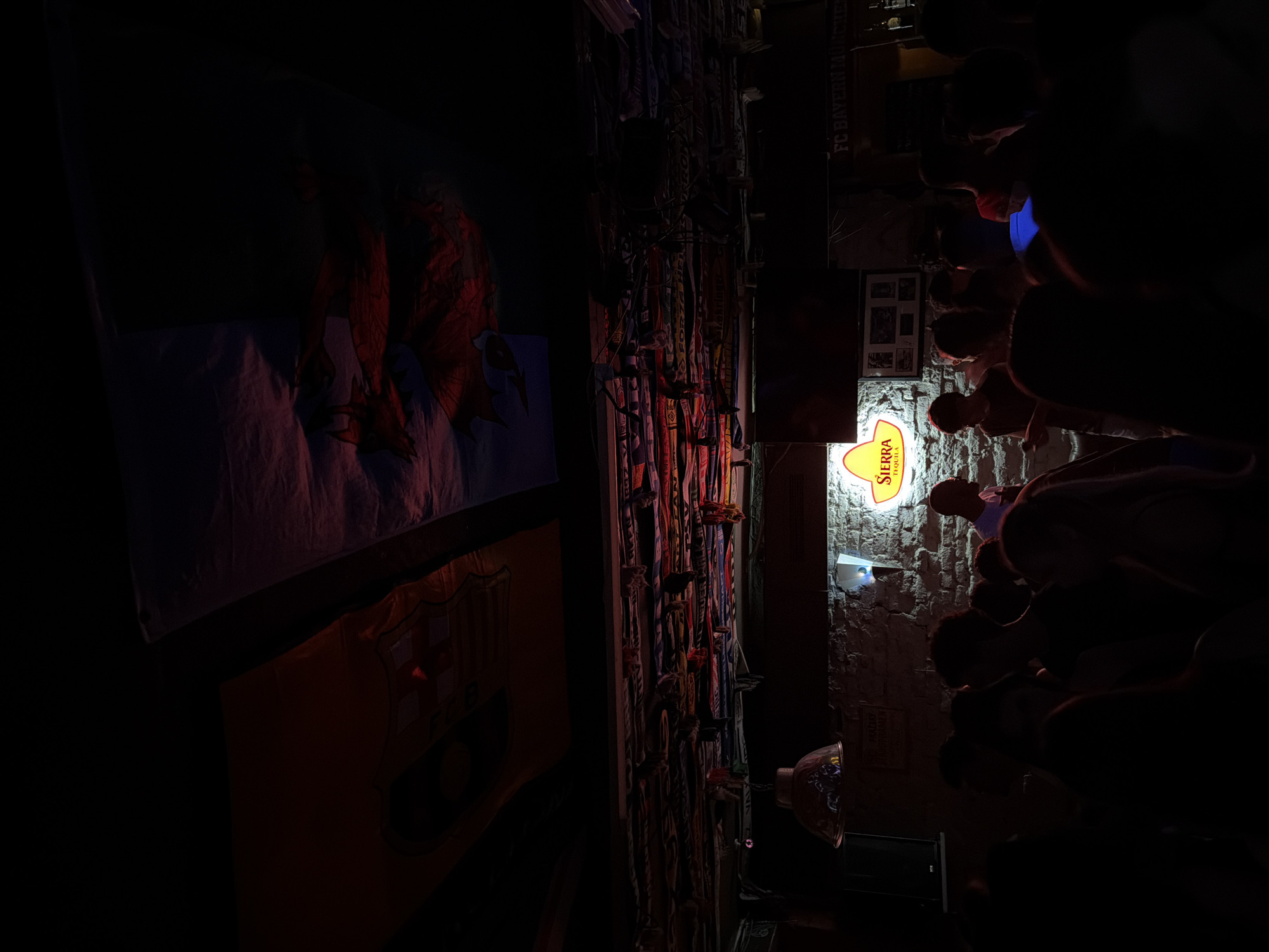 The image shows the interior of a bar or pub in Plovdiv, Bulgaria. On the left, there are flags, including one possibly associated with a sports team (FC Barcelona). A collection of sports scarves is affixed to the wall. On the right, a Sierra Tequila sign illuminates a roughly textured stone wall, behind a row of people. A projection is visible on the wall below the Sierra Tequila sign. A FC Bayern München banner is visible above the people. The lighting in the establishment is dim.