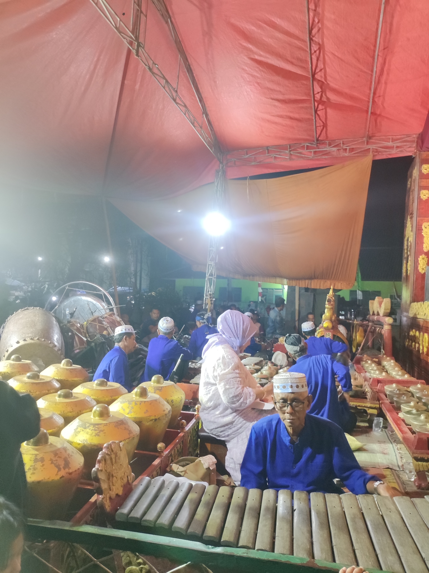 In Panongan, Indonesia, a group of people are gathered around traditional gamelan instruments, likely for a performance or rehearsal. The scene is illuminated by artificial lighting under a large red tent structure supported by metal scaffolding. In the foreground, an elderly man wearing glasses, a blue long-sleeved shirt, and a white peci (cap) is seated, appearing to be a musician. He is positioned in front of a xylophone-like instrument. Behind him, other individuals are also engaged with various percussion instruments, including large gongs and smaller tuned metal bars. Some are dressed in similar blue attire and peci caps, suggesting a uniform. A woman in a light-colored hijab and dress is visible further back, interacting with the instruments. The overall atmosphere suggests a cultural event or musical gathering in an outdoor or semi-outdoor setting.