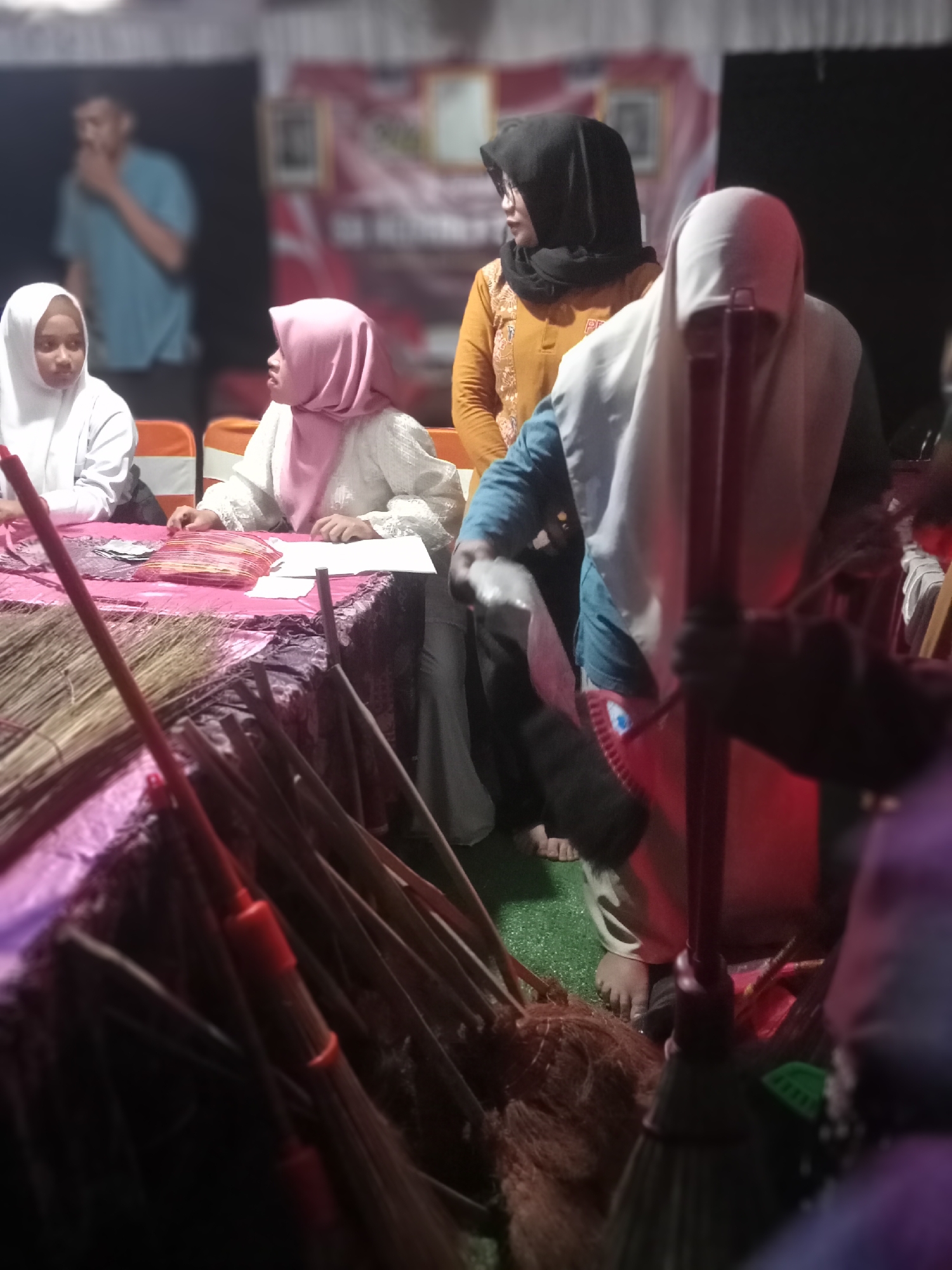 In Pule, Indonesia, a low-light indoor scene captures a group of individuals gathered around a table covered with a pink patterned tablecloth. Two young females wearing hijabs, one in white and another in pink, are seated at the table. The female in pink appears to be interacting with items on the table, possibly papers or fabric. To their right, a woman in a dark hijab and an orange top is partially visible, and another woman in a white hijab and a blue top is positioned further back. In the foreground, several brooms are arranged vertically, leaning against the table. The environment appears to be some kind of community event or gathering, with a blurred banner visible in the background.