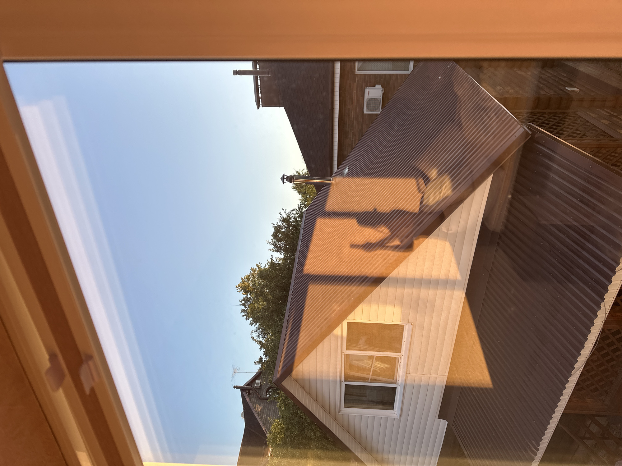 The image depicts a view through a window of suburban rooftops in Nizhny Novgorod, Russia, bathed in the warm light of sunset. The foreground shows a window frame with a wooden interior. The sky is a clear, pale blue, indicating late afternoon or early evening.
Dominating the scene are several house roofs. On the left, a gabled roof with white siding is visible, featuring a window. The large, sloped roof of another house, covered in dark brown corrugated metal, extends across the center and right of the frame, casting long shadows. A portion of a third roof, also dark brown and likely shingled, is visible behind the corrugated one. A chimney extends upwards from this roof. On the left side, a taller, older house with a grey, shingled roof and a prominent antenna is partially obscured by trees. An outdoor air conditioning unit is mounted on the wall of one of the brick houses.
The sunlight casts dramatic shadows and highlights across the scene, accentuating the textures of the roofs and siding. The overall impression is one of a peaceful, residential setting at the end of the day. There are no people or animals visible in the image.