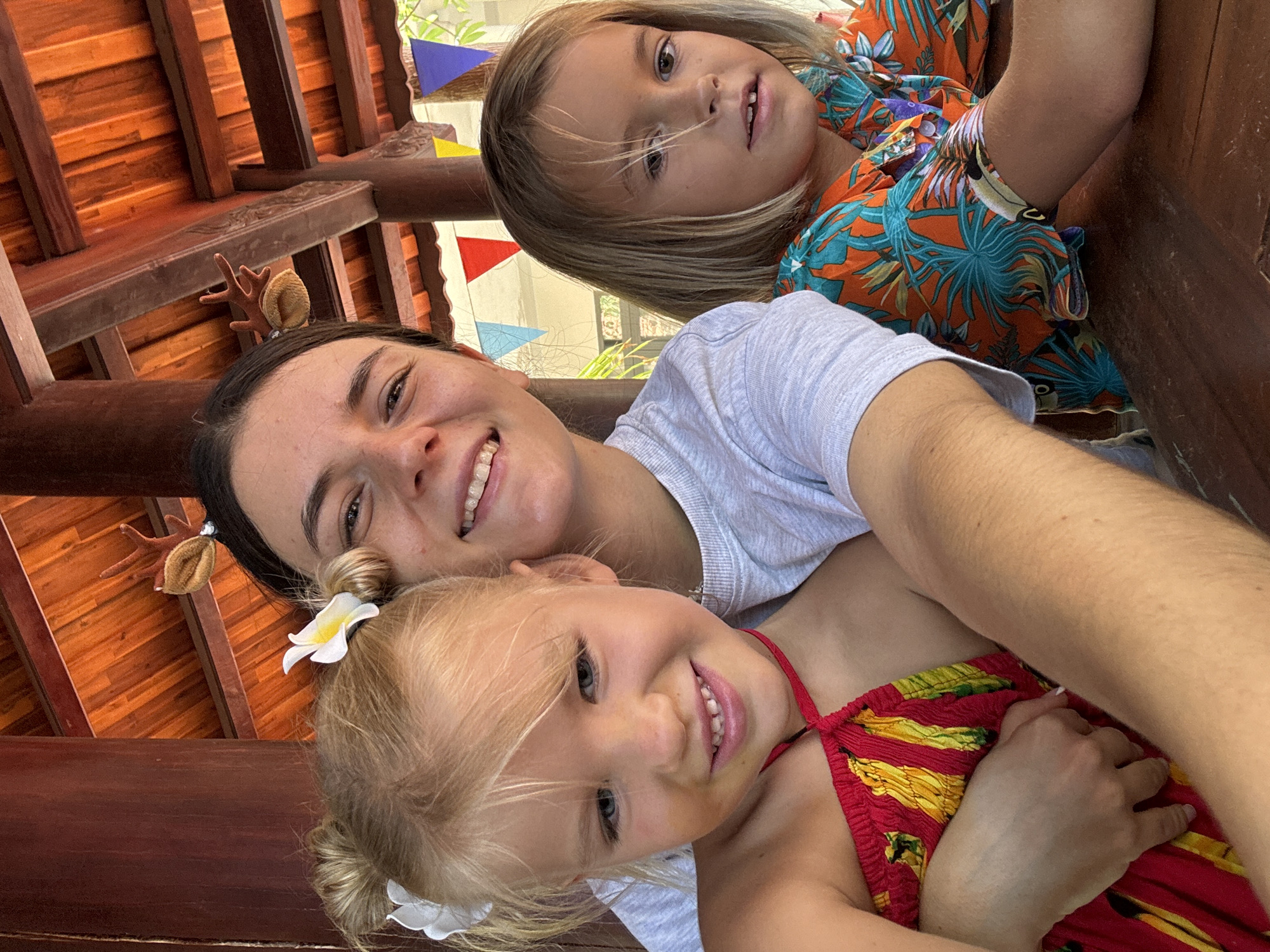 The image is a low-angle shot of a woman and two children. The woman is positioned in the middle, with a child with blonde hair and a white flower in their hair below her. Above the woman is a second child with blonde hair. All three individuals are smiling and looking toward the camera.
The woman has dark hair styled with decorative accessories and is wearing a light gray t-shirt. The child below her is wearing a red and yellow printed top. The second child above the woman is wearing an orange and blue tropical-print shirt.
The background includes a wooden structure, possibly part of a building or gazebo, as well as colorful triangular pennants strung overhead. The location is Mũi Né, Vietnam, implied by the tropical clothing and possible building style.