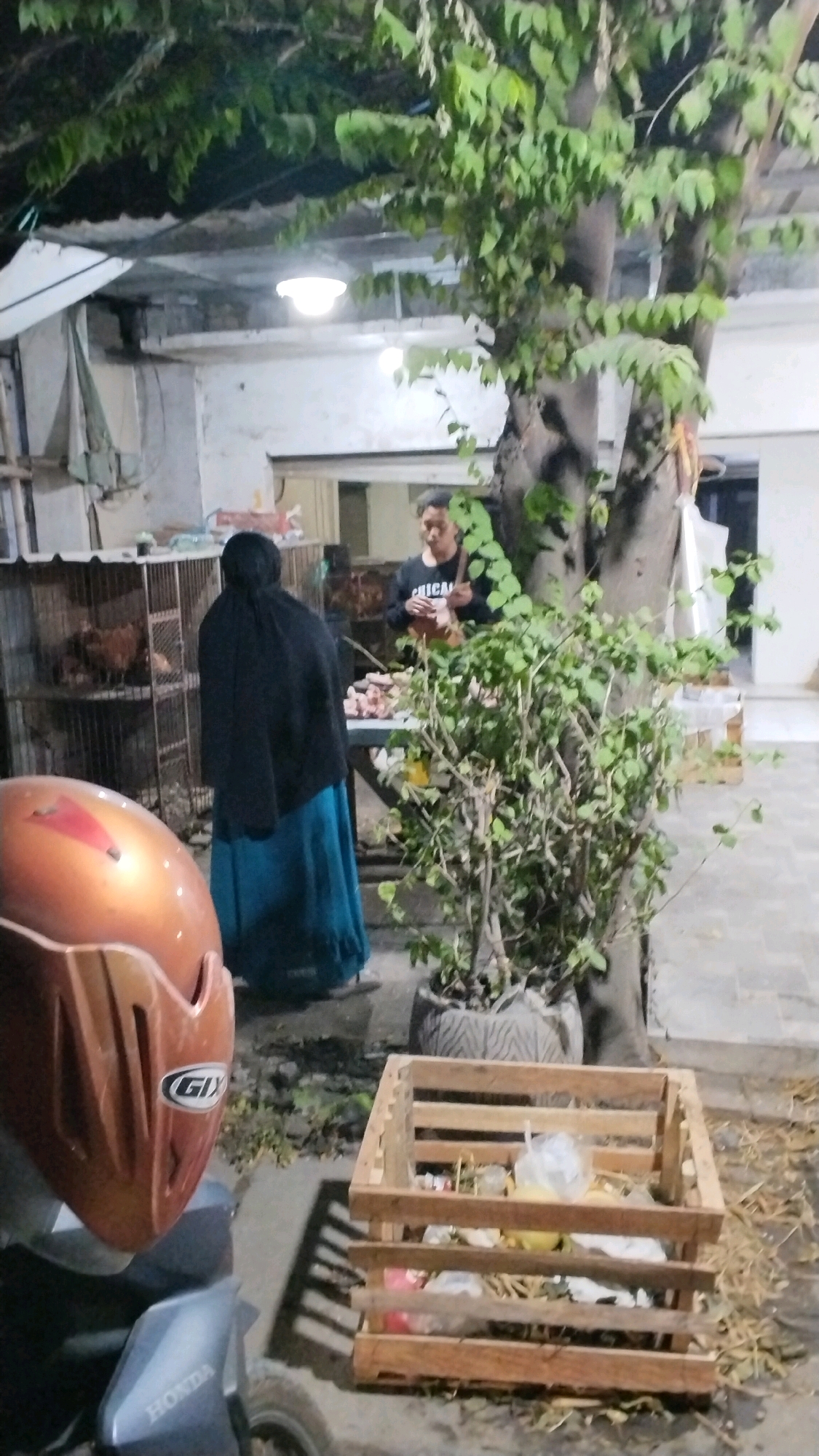 The image depicts an outdoor scene, likely in Canggu, Indonesia, during nighttime. In the foreground, a motorcycle helmet, predominantly orange with a grey visor, is visible on the left. To the right of the helmet, a wooden crate filled with various discarded items, including plastic bags and what appears to be food waste, is situated on the ground.
In the mid-ground, a person wearing a black headscarf and a blue skirt stands facing away from the camera, next to a cage containing chickens. To their right, a table is laden with what seems to be raw meat. A man wearing a black sweatshirt with the word "CHICAGO" is standing behind the table, appearing to engage with the meat. A large tree with dense green foliage dominates the right side of the image. The environment suggests an informal market or a residential area with agricultural elements. The lighting is dim, originating from overhead lamps.