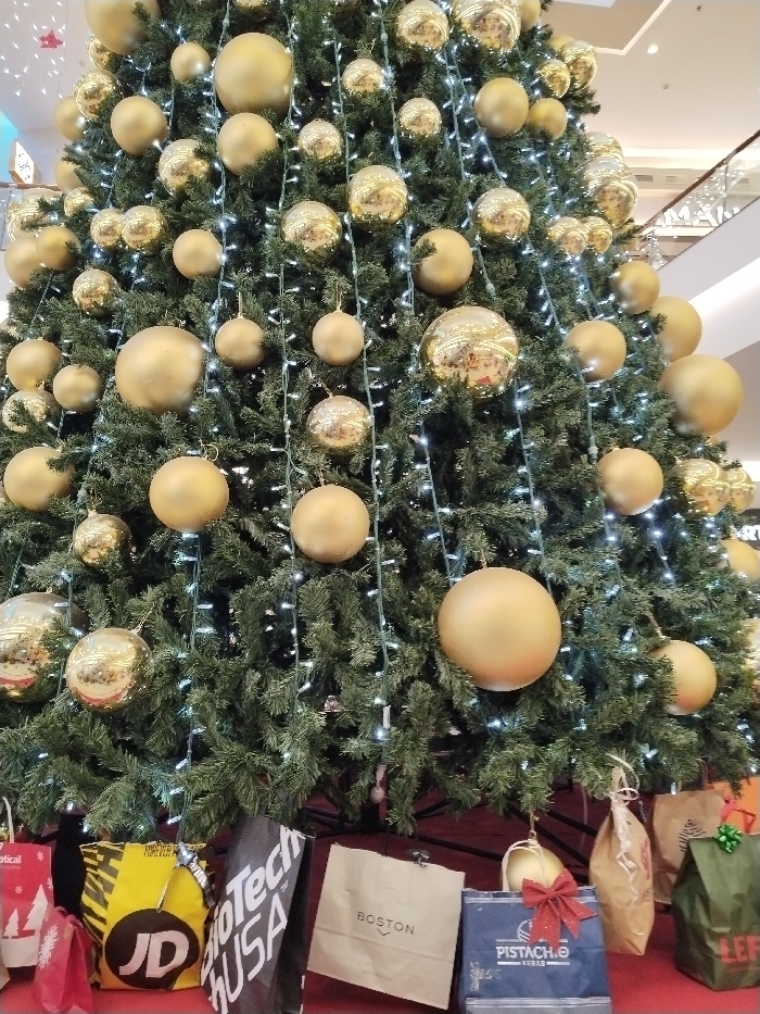 This image depicts a large, artificially constructed Christmas tree situated within the C.C. Dos Mares shopping center in San Javier, Spain. The tree features dense, dark green faux foliage and is extensively decorated with numerous spherical ornaments, uniformly gold in color. These ornaments vary in size and possess either a matte or highly reflective finish; the reflective ornaments display distorted reflections of the surrounding indoor environment. Strands of small, white LED lights are interwoven throughout the branches. The base of the tree is covered by a red material, upon which several shopping bags are arranged. These bags include identifiable branding such as "JD," "BioTech USA," "BOSTON," and "PISTACH.O." The background reveals elements consistent with a multi-level shopping mall interior, including upper-level architectural features and a partial view of an escalator, under bright overhead lighting. No individuals are visible within the scene.
