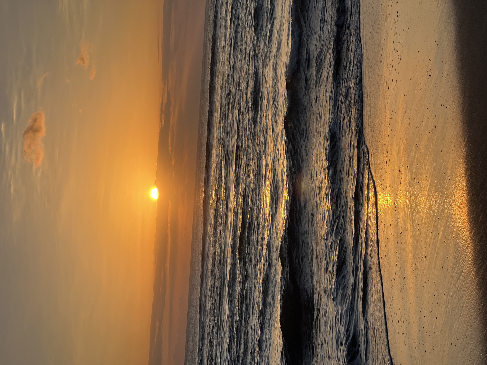 The image depicts a serene ocean sunset at Kuta Beach, Indonesia. The sun, a bright orb of orange, is partially obscured by soft, wispy clouds, casting a warm, golden glow across the sky and water. Gentle waves roll towards the sandy shore, their surfaces shimmering with the reflected sunlight. The foreground is dominated by wet sand, glistening with the receding tide and speckled with small, dark bubbles. The overall impression is one of tranquility and natural beauty, characteristic of a peaceful evening on a tropical beach. There are no people or specific objects visible in the frame, emphasizing the natural landscape.