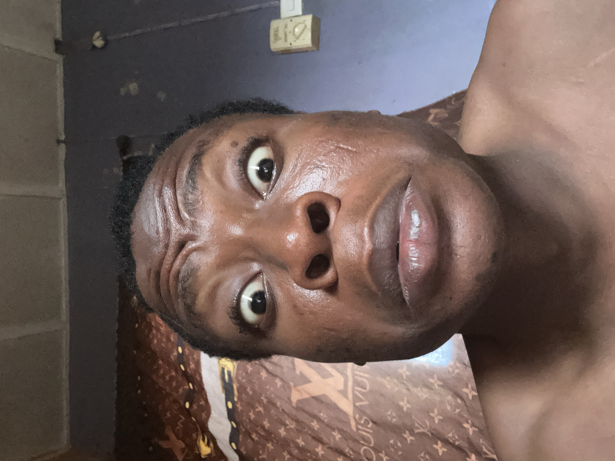 A close-up, upward-facing portrait of a dark-skinned person with a visibly furrowed brow. The person's eyes are wide, suggesting surprise or alertness. The background includes a gray wall with a small, rectangular object attached near the top. Part of a bed with a Louis Vuitton-patterned covering is visible behind the person. The location is not discernible from the image.