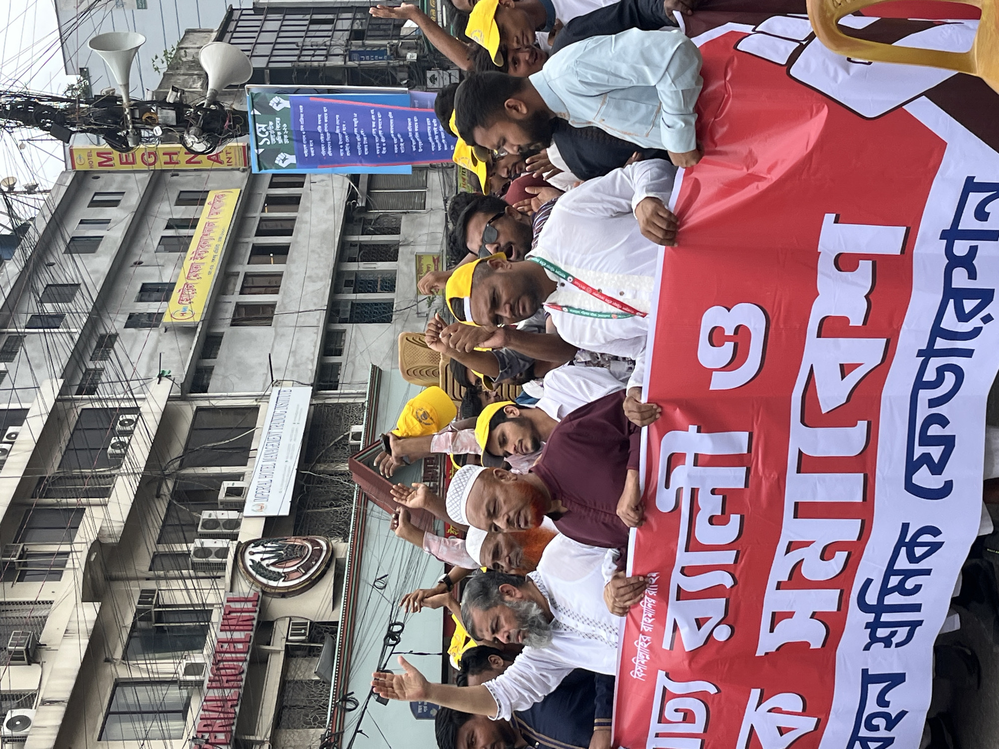 A street-level photograph captures a crowd of men participating in a rally or demonstration in Dhaka, Bangladesh. The subjects are mostly young to middle-aged men, many wearing traditional attire such as kurtas or panjabis, with some sporting yellow caps. They are holding aloft a large red and white banner, which dominates the foreground.
The banner's Bengali text clearly indicates the event: "১মে আন্তর্জাতিক শ্রমিক দিবসের র্যালী ও সমাবেশ" (May 1st International Workers' Day Rally and Assembly). Smaller text on the banner includes "বিসমিল্লাহির রাহমানির রাহিম" (In the name of Allah, the Most Gracious, the Most Merciful) and "বহন শ্রমিক ফেডারেশন" (Bahan Sromik Federation / Transport Workers' Federation), identifying the organizing body. Some individuals in the crowd have their hands raised in what appears to be a gesture of solidarity or participation.
In the background, multiple buildings line the street, featuring various signs. A prominent yellow banner reads "HOTEL MEGHNA INT" (মেঘনা হোটেল ইন্টাঃ). Another blue banner, partly visible, displays Bengali text with a raised fist logo, proclaiming "সংগঠিত হও, প্রতিরোধ করো" (Be organized, resist), a common workers' solidarity slogan. Higher up, a white sign identifies "IMPERIAL HOTEL MANAGEMENT TRAINING INSTITUTE". The urban setting is characterized by numerous tangled electrical wires crisscrossing overhead and several loudspeakers attached to poles. The lighting suggests it is daytime, possibly overcast, creating a soft, diffuse illumination. The overall scene depicts a vibrant street protest or gathering for International Workers' Day.