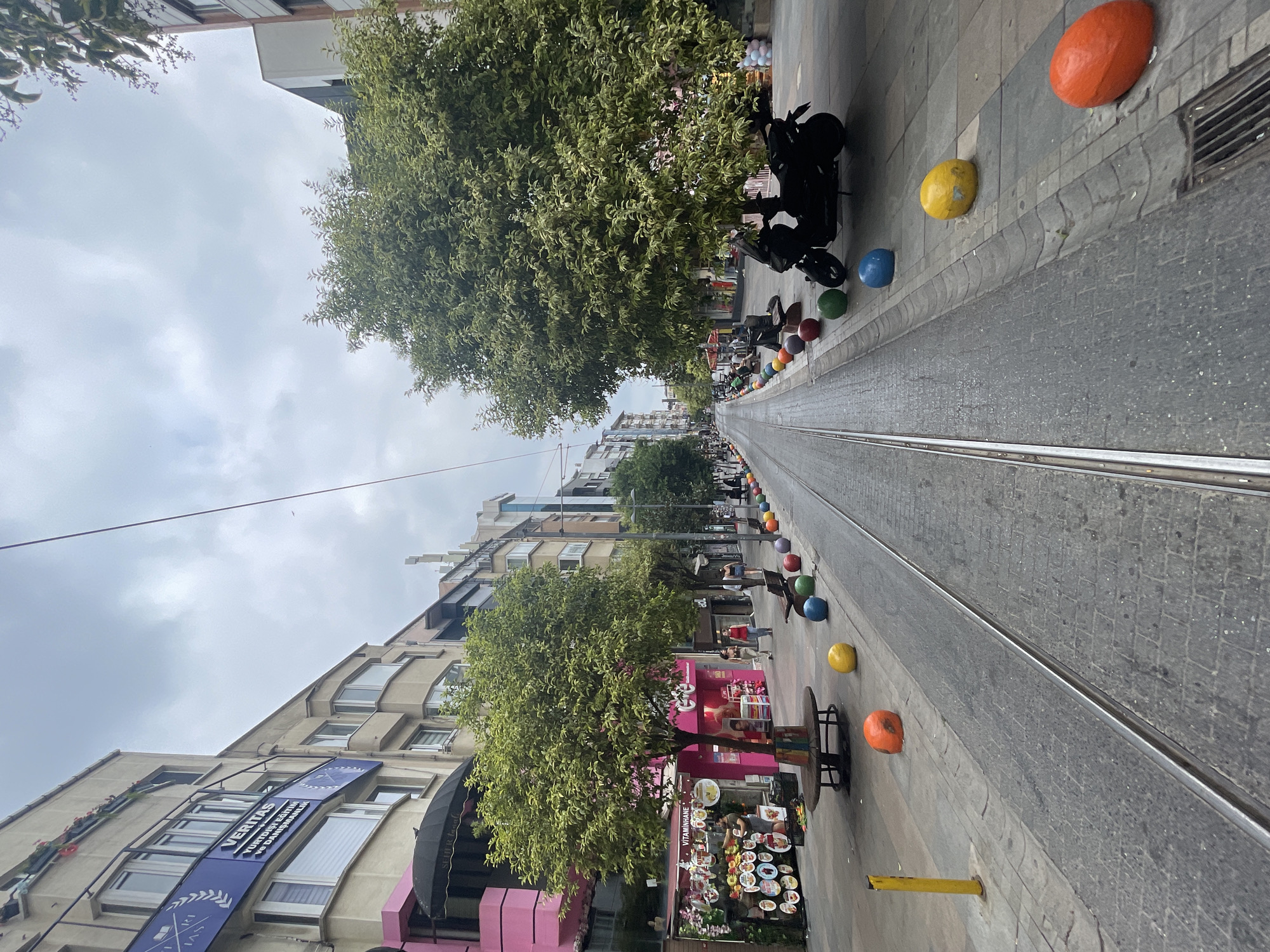 The image captures a street scene in Kadıköy, Turkey, characterized by tram tracks running down the center of the road. On either side of the tracks, colorful, rounded bollards are interspersed with trees and benches. Buildings line the street, featuring storefronts at ground level and windows on upper floors, with some displaying signage. A black scooter is parked on the right side of the road, near some of the bollards and trees. A few individuals are visible walking on the sidewalks and browsing the shops. The sky is overcast. The scene appears to be a pedestrian-friendly commercial street with elements of urban greenery and public seating.