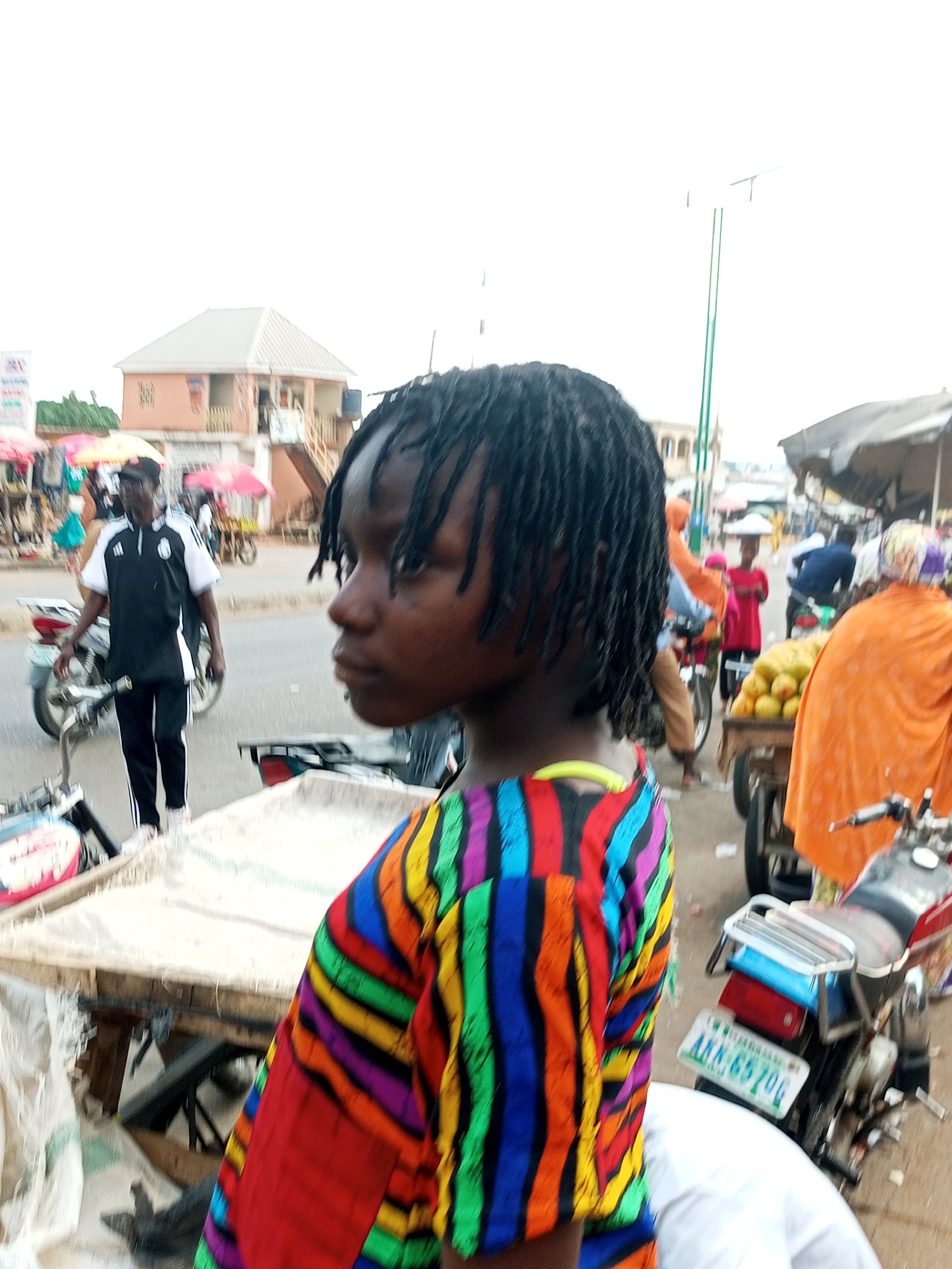 The image depicts a bustling outdoor market scene in the city of Unknown, Unknown. In the foreground, a young individual with dark dreadlocks is shown in profile, wearing a vibrantly colored, striped shirt with patterns of red, yellow, blue, green, and purple. To the left of the foreground subject, a man in a black and white athletic-style shirt and black pants stands near a motorcycle. In the middle ground, other individuals are visible, including one wearing a bright orange garment and another in a pink shirt, seemingly involved in the market's commerce. A cart laden with yellow fruits, possibly mangoes, is also present. Several motorcycles are parked or in motion within the scene, indicating transportation. The background shows buildings and stalls, suggesting the market is set up along a street. The sky is overcast.