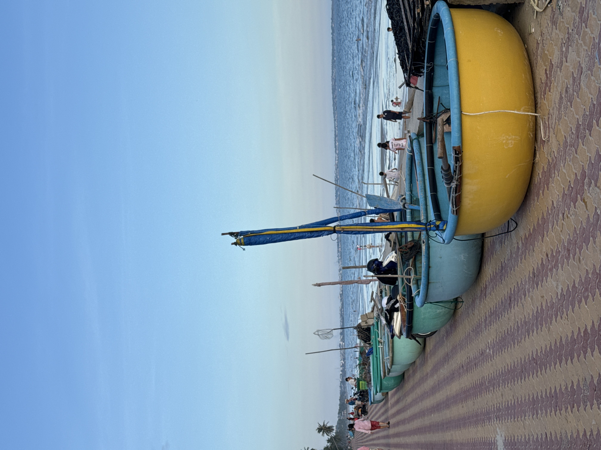 The image depicts a seaside scene in Mũi Né, Vietnam. A paved walkway, laid with reddish-brown and tan interlocking bricks, runs along the left side of the frame. A line of round, basket-like boats, painted in various shades of green and blue, are arranged on the sand next to the walkway. The boat in the foreground is bright yellow with a blue rim. Several people are visible walking on the walkway and near the boats, dressed in casual attire. Beyond the boats, the ocean stretches out, with gentle waves lapping at the shore. Several people are in the water, and a few appear to be playing on the beach. In the distance, a hazy shoreline with buildings and palm trees is visible under a clear, pale blue sky. The lighting suggests it is late afternoon or early evening.