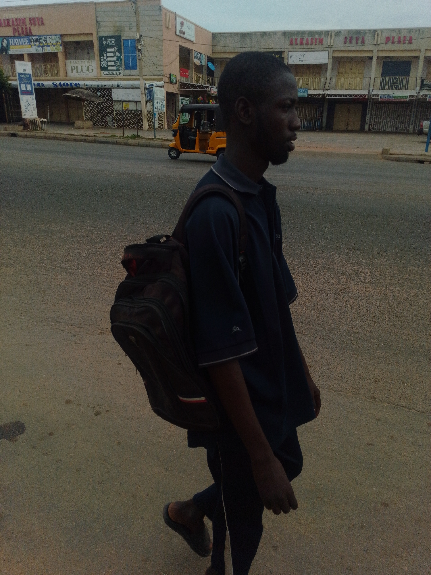 A medium shot captures a young man walking on an asphalt road in Gombe, Nigeria. He is seen from the chest up on the right side of the frame, with his body turned to the right, facing away from the camera. The man has dark skin, short black hair, and a trimmed beard. He is wearing a dark blue polo shirt with white trim around the collar and sleeve edges, and dark trousers. A dark-colored backpack with red and white accents is slung over his left shoulder. The background reveals a street scene with commercial buildings and a yellow three-wheeled auto-rickshaw, known locally as a "Keke Napep," moving away from the camera. The buildings are painted in light colors and feature signage, including "ALKASIM SUYA PLAZA," "FASHION PLUG," and "HAWEE." The sky is overcast, suggesting a muted lighting condition. The road surface shows some minor stains.