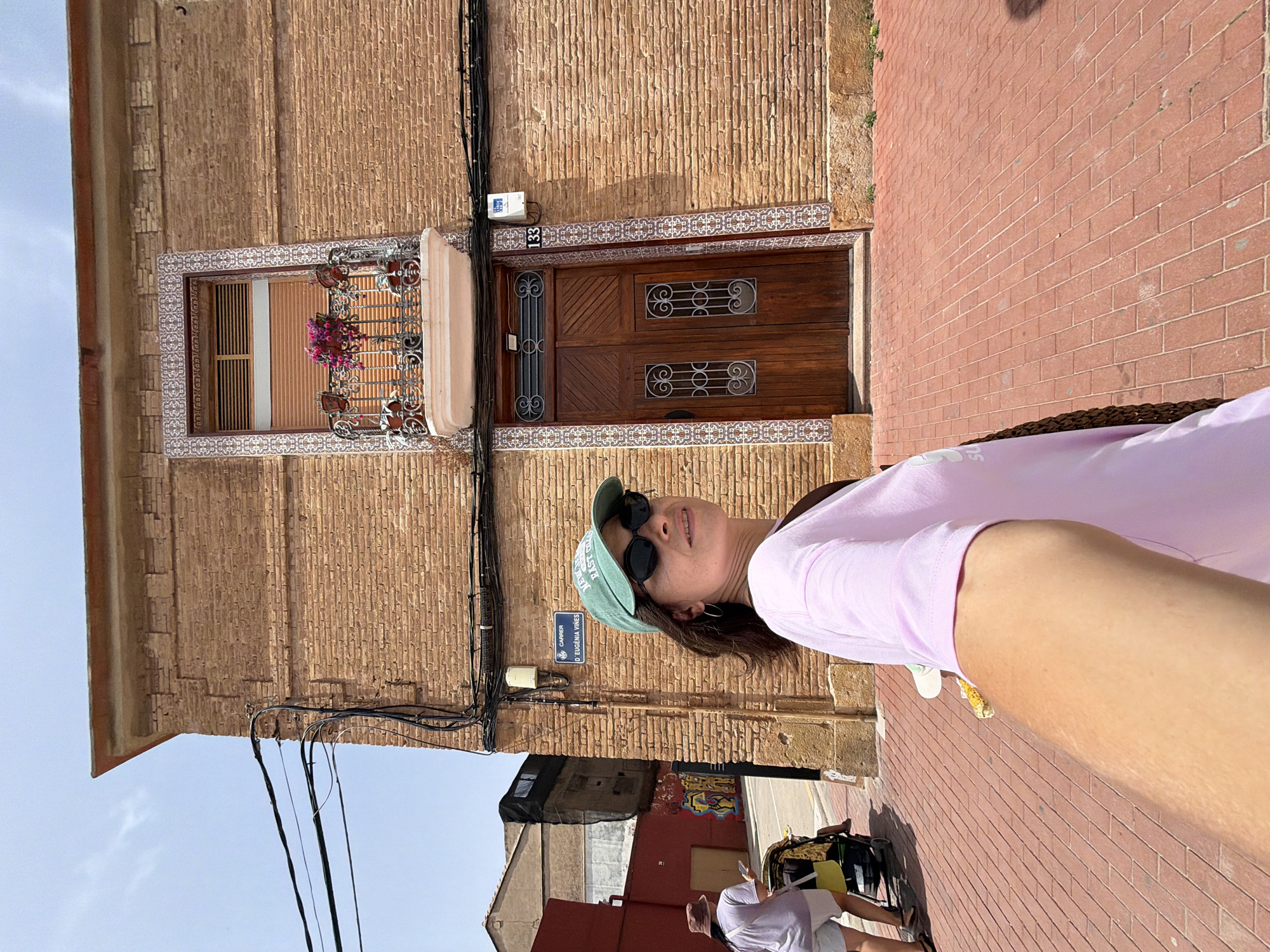 A woman wearing a pink t-shirt, sunglasses, and a green baseball cap poses for a selfie outdoors. She is standing on a brick-paved sidewalk next to a brick building. The building has a wooden door with decorative metal accents and a window with a wrought-iron grate and potted flowers. The time of day appears to be sunny, and the weather is clear. There is a street sign that reads "Carrer D'Eugenia VInes." In the background, other buildings and people are visible, suggesting an urban environment. The woman has a relaxed expression, and the overall mood of the image is casual and summery.