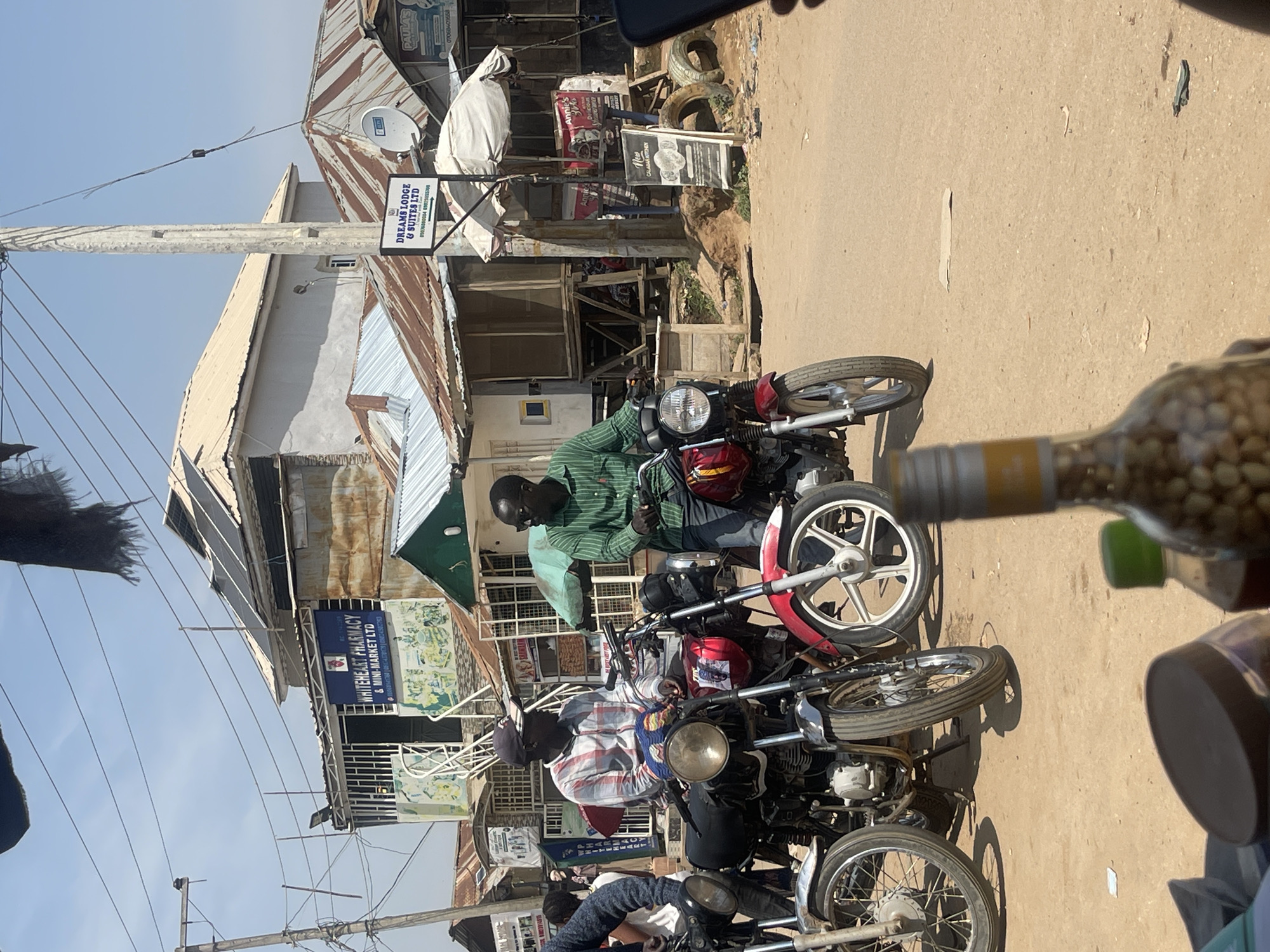 The image captures a street scene in Abuja, Nigeria, likely a bustling market or commercial area. Two men are seated on motorcycles in the foreground, facing each other and engaged in conversation. The man on the left wears a checkered shirt and a baseball cap, while the man on the right is dressed in a green striped shirt. Both motorcycles are positioned on a dusty, unpaved road.
Behind the motorcycles, a row of shops and buildings lines the street. One prominent sign reads "WHITEHEART PHARMACY & MINI-MARKET LTD". Another sign indicates "DREAMS LODGE & SUITES LTD". The architecture suggests a mix of modern and older structures, with corrugated iron roofs visible on some buildings. Solar panels are also noticeable on the roof of one building, indicating a potential adoption of renewable energy.
The environment appears sunny and dry, with clear skies overhead. A few other individuals are visible in the background, along with more motorcycles, suggesting vehicular activity and pedestrian traffic. The overall impression is one of a typical day in a Nigerian urban setting, with commerce and daily life unfolding on the streets. The presence of bottles and other items in the very foreground suggests that the photo was taken from a vendor's stall or a similar vantage point.