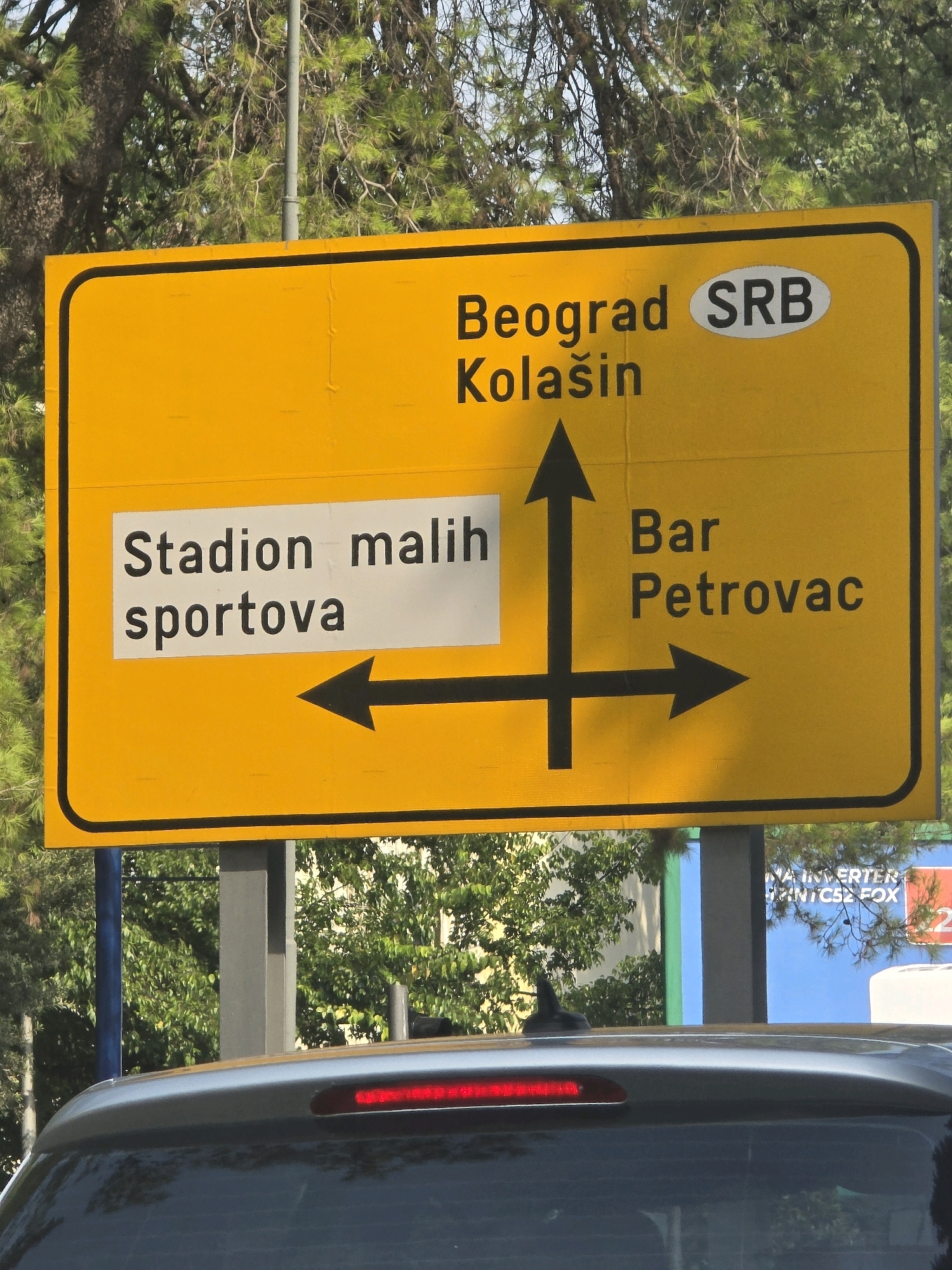 A bright yellow, rectangular road sign with black lettering and directional arrows is mounted on a gray pole behind a silver car. The sign displays multiple destinations. At the top, "Beograd" is written with "SRB" in an oval to its right. Below "Beograd" is "Kolašin". To the left, a white rectangular section on the yellow sign reads "Stadion malih" above "sportova" with a black arrow pointing left. To the right of the center, "Bar" is written above "Petrovac" with a black arrow pointing right. The background consists of green trees and a blue sky. The car in the foreground is partially obscuring the bottom of the sign and the pole. A red brake light is visible on the car's rear. The scene appears to be a roadside in Podgorica, Montenegro, based on the destinations listed.