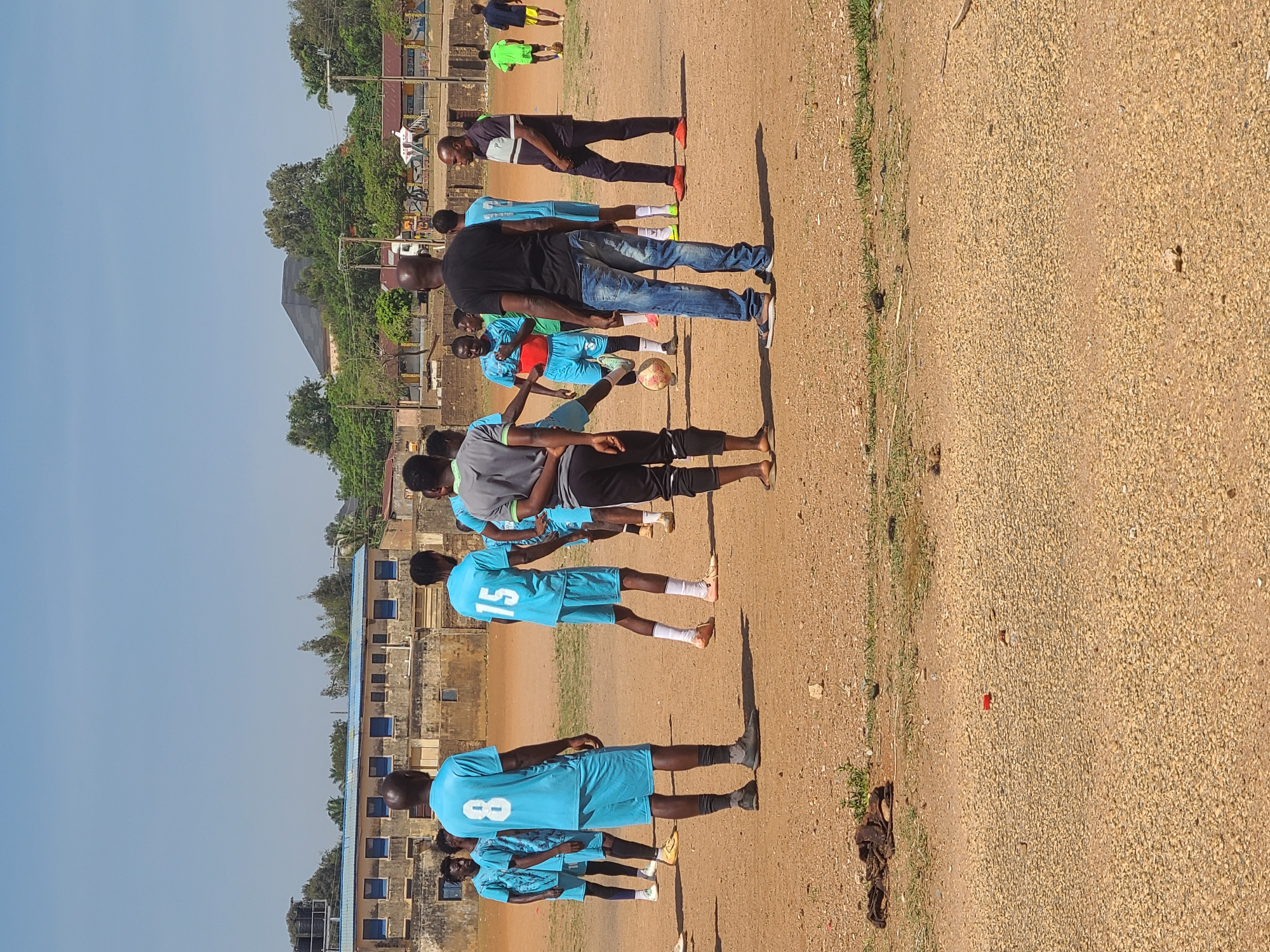 The image captures an outdoor scene featuring a group of individuals, primarily young males, gathered on a dusty, unpaved field under a clear blue sky. The subjects appear to be involved in a soccer practice or an informal game. Several young men are wearing light blue athletic jerseys, with the numbers "8" and "15" prominently visible on two of them. A soccer ball is visible on the ground near the center of the group.
Two adult males are also present: one in a black t-shirt and blue jeans stands among the younger players, while another, wearing a striped polo shirt and dark trousers, stands further back. The ground is predominantly reddish-brown dirt and gravel, with sparse patches of green grass along the edges.
The clear blue sky and prominent shadows indicate bright, sunny weather, likely during the morning or afternoon. In the background, various buildings are visible, some appearing to be residential or communal structures with multiple windows. One specific building features numerous small, rectangular windows with blue coverings. Lush green trees line the horizon behind the buildings. The overall setting suggests a community sports activity in a semi-urban or rural environment.