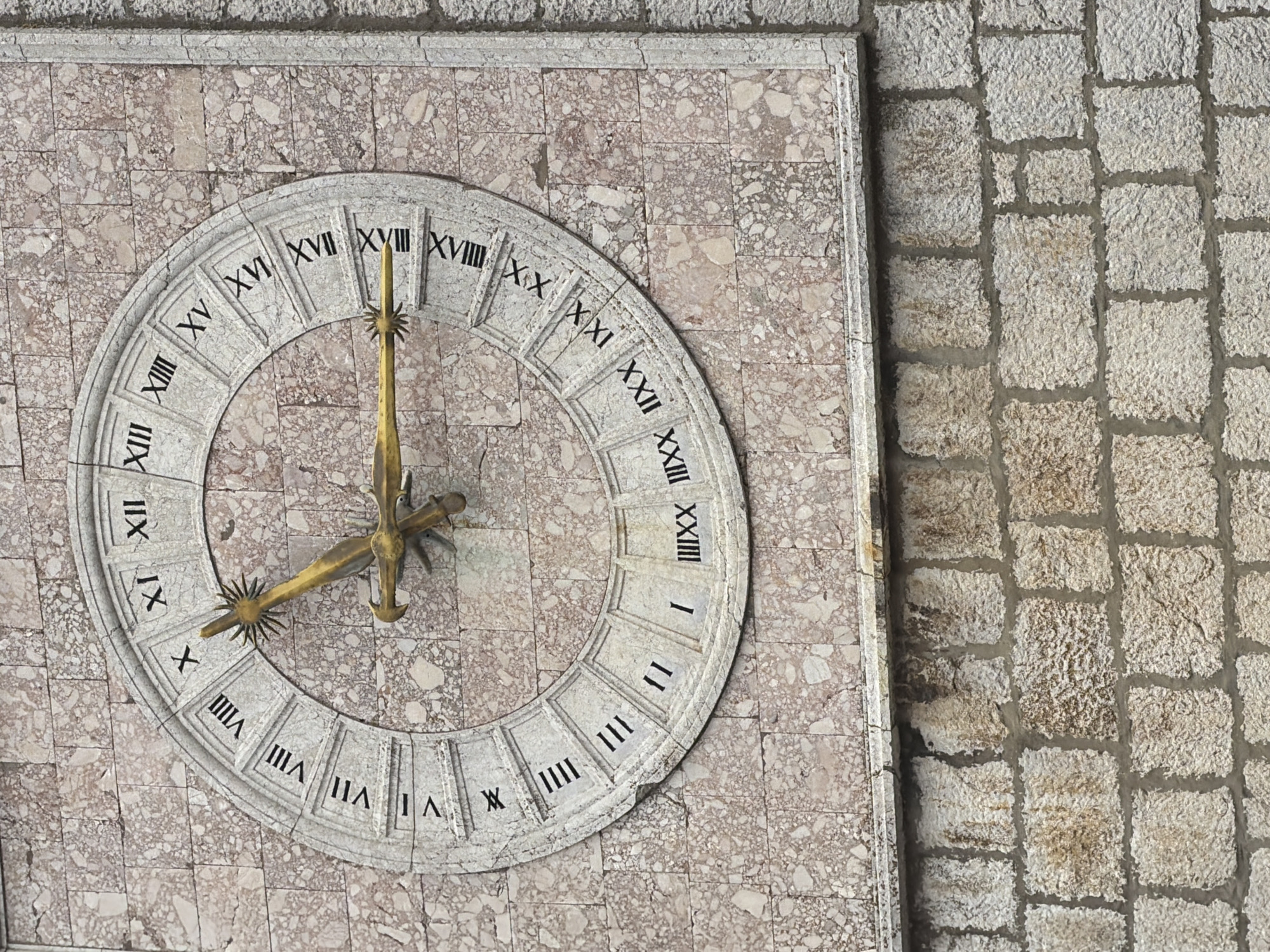This is a close-up, eye-level, medium shot of a large, ornate clock face mounted on a stone wall in the city of Krk, Croatia. The clock face is circular and made of a light-colored material, possibly stone or marble, with Roman numerals indicating the hours. The numerals range from I to XXIV, suggesting a 24-hour clock. The clock has two golden hands; the hour hand is shorter and thicker, while the minute hand is longer and thinner, both featuring decorative tips. The clock is set against a wall constructed of rectangular stone blocks of varying sizes, with a reddish-pink marbled pattern visible behind the clock and a rougher, lighter-colored stone below. The clock is positioned on a building, as indicated by the architectural style of the surrounding stonework. The image captures the detail of the clock's Roman numerals and the texture of the stone.