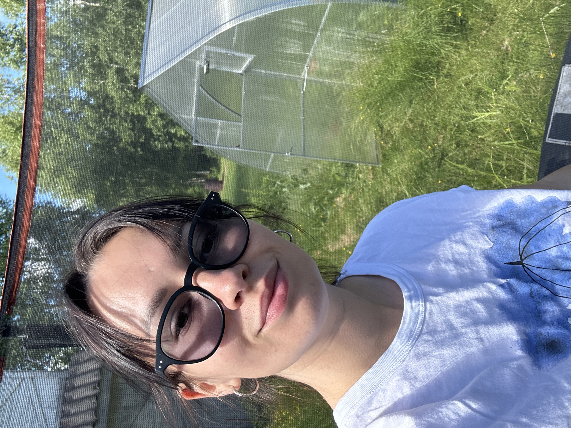 The image is a selfie taken outdoors, likely in the city of Novozhilovo, Russia, on a sunny day. A young woman with dark hair and glasses is in the foreground, smiling at the camera. She is wearing a white t-shirt with a blue abstract design. Behind her, to the right, is a small greenhouse with a transparent cover. The background consists of lush green grass, some scattered yellow wildflowers, and trees. A portion of a building with a tiled roof is visible to the left, and a black net structure with a red trim is present at the top of the frame, possibly part of a trampoline or a protective enclosure. The overall impression is a casual outdoor scene in a rural or suburban setting.