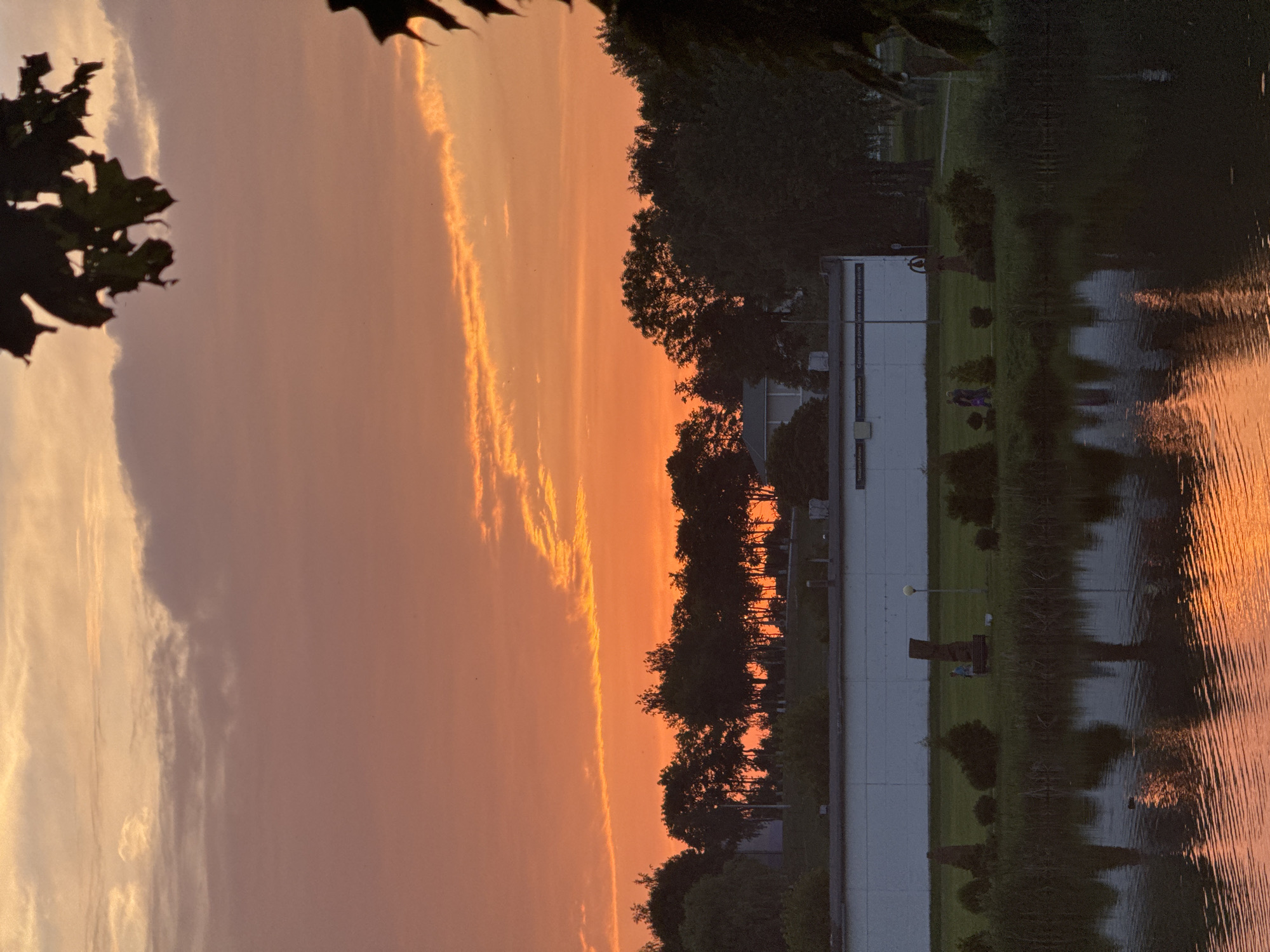 A sunset over a body of water, possibly a pond or lake, in Nesvizh, Belarus. The sky is filled with vibrant shades of orange and yellow, with streaks of light clouds. In the foreground, the water reflects the colors of the sunset, creating a shimmering effect. Along the bank, there are trees and grass, and a white building is visible in the background. There are a few people walking on the grass near some sculptures. The overall scene is serene and picturesque.