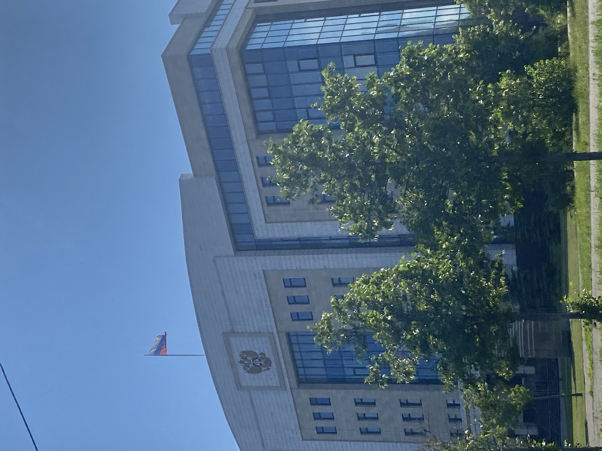 The image shows a large, modern building with a flag of Russia flying from a flagpole on its roof. The building appears to be made of stone and glass, with several stories and many windows. In front of the building, there are several trees with green leaves. The sky is blue and clear, indicating a sunny day. Based on the Russian flag and the architectural style, this building is likely a government institution located in the city of Kazan, Russia.