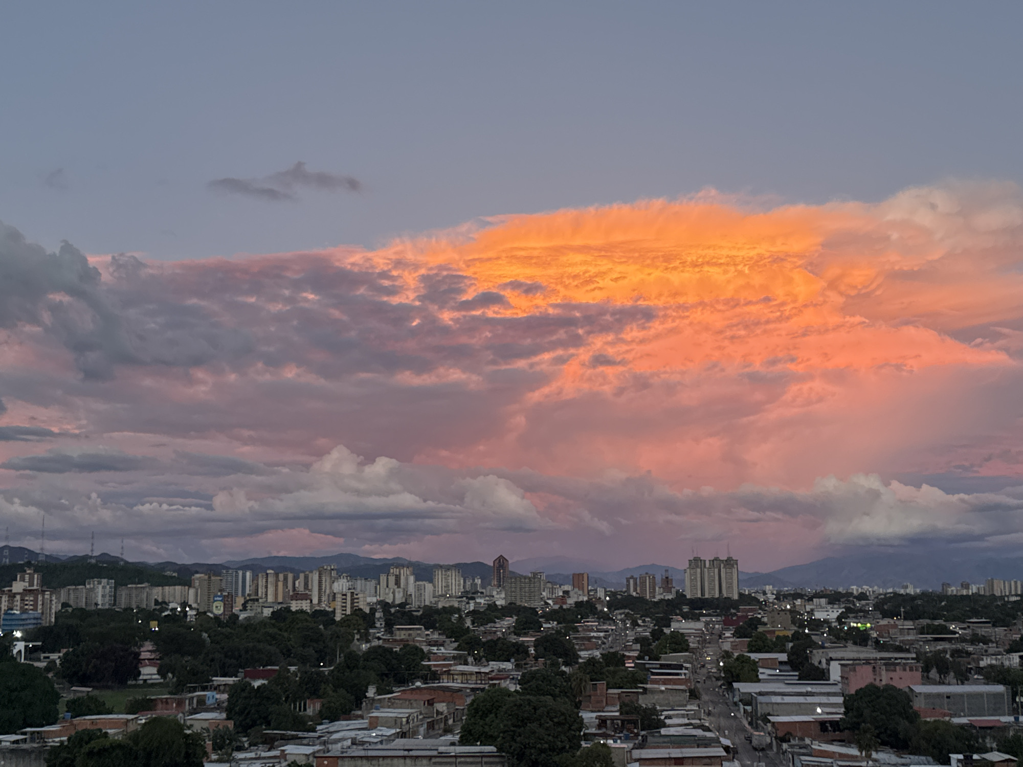 The image depicts a cityscape of Maracay, Venezuela, under a dynamic sky at twilight or dawn. The lower part of the image shows residential and commercial buildings, many of which are low-rise with flat or slightly sloping roofs. Patches of trees are interspersed among the buildings, suggesting urban greenery.
In the mid-ground, there is a line of taller buildings forming the city's skyline, with a few high-rise structures visible against the backdrop of distant mountains.
The sky dominates the upper portion of the image, filled with a complex pattern of clouds. The most striking feature is a large, brightly lit, orange-colored cloud formation. Other clouds range in color from grey to pink to white, adding depth and texture to the scene. The overall lighting suggests either a sunset or sunrise, with the sun casting a warm glow on the clouds.