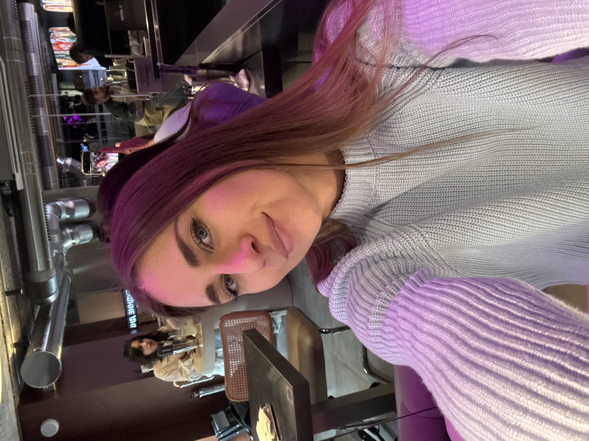 The image captures a selfie of a young woman inside a modern, dimly lit bar or restaurant in Moscow, Russia. The woman, with long reddish-brown hair (appearing magenta due to lighting) and light blue eyes, is in the foreground, wearing a light grey/blue ribbed knit sweater and subtly smiling while looking directly at the camera. Her right arm and shoulder are visible, extending towards the camera.
In the background, the establishment features an industrial-chic interior with exposed metal ventilation ducts running across the ceiling. To the left, a woman with dark hair, dressed in a light-colored top, is seated at a round table, holding what appears to be a cocktail shaker or a tall glass. Near her, a small square table with a dark top and a modern chair featuring a cane back and brown seat is visible; a stack of light-colored square items (possibly coasters or napkins) rests on this table. A neon sign with Cyrillic text, reading "И НА ЗНАНИЕ" (And on Knowledge), is visible on a wall in the mid-background.
To the right, a bar area is present with a black counter. A bearded man in a green and beige top is seated at the bar, while another individual is partially visible behind the counter. Various bottles, bar equipment, and computer monitors are positioned on and behind the bar. The overall lighting is artificial and subdued, with strong pink/magenta lighting illuminating the main subject's face and hair, and other colorful ambient lights reflecting in the background. The specific name or precise address of the establishment is not discernible from the visual information provided, but it is characteristic of a contemporary commercial venue in Moscow.