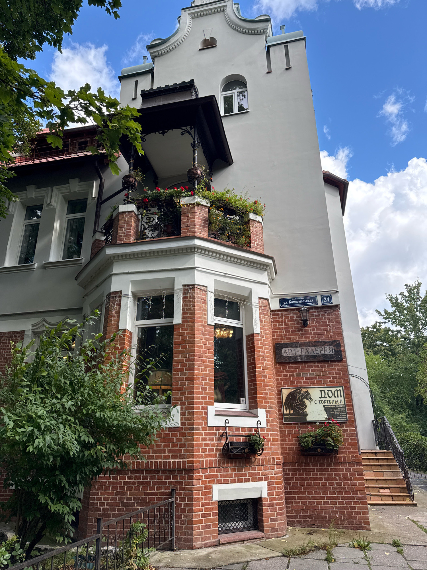 A slightly low-angle shot captures the facade of a red brick and grey stucco building in Kaliningrad, Russia, under a bright, partly cloudy sky. The building features a prominent corner bay with red brickwork, large windows with white trim, and a second-story balcony adorned with potted red flowers and greenery. Above the bay, a sign reads "ул. Комсомольская 24" (Komsomolskaya Street 24). Below the balcony, a sign with dark lettering on a light background reads "АРТ-ГАЛЕРЕЯ" (Art Gallery). Further down on the brickwork, a framed sign depicts a mythological creature and text that appears to include "ДОМ С ТОРГОВЛЕЙ" (House with Trade). A set of outdoor stairs with a black metal railing ascends the right side of the building. Lush green trees and foliage frame the left side of the image, casting shadows on the building and the paved walkway. No people are visible.