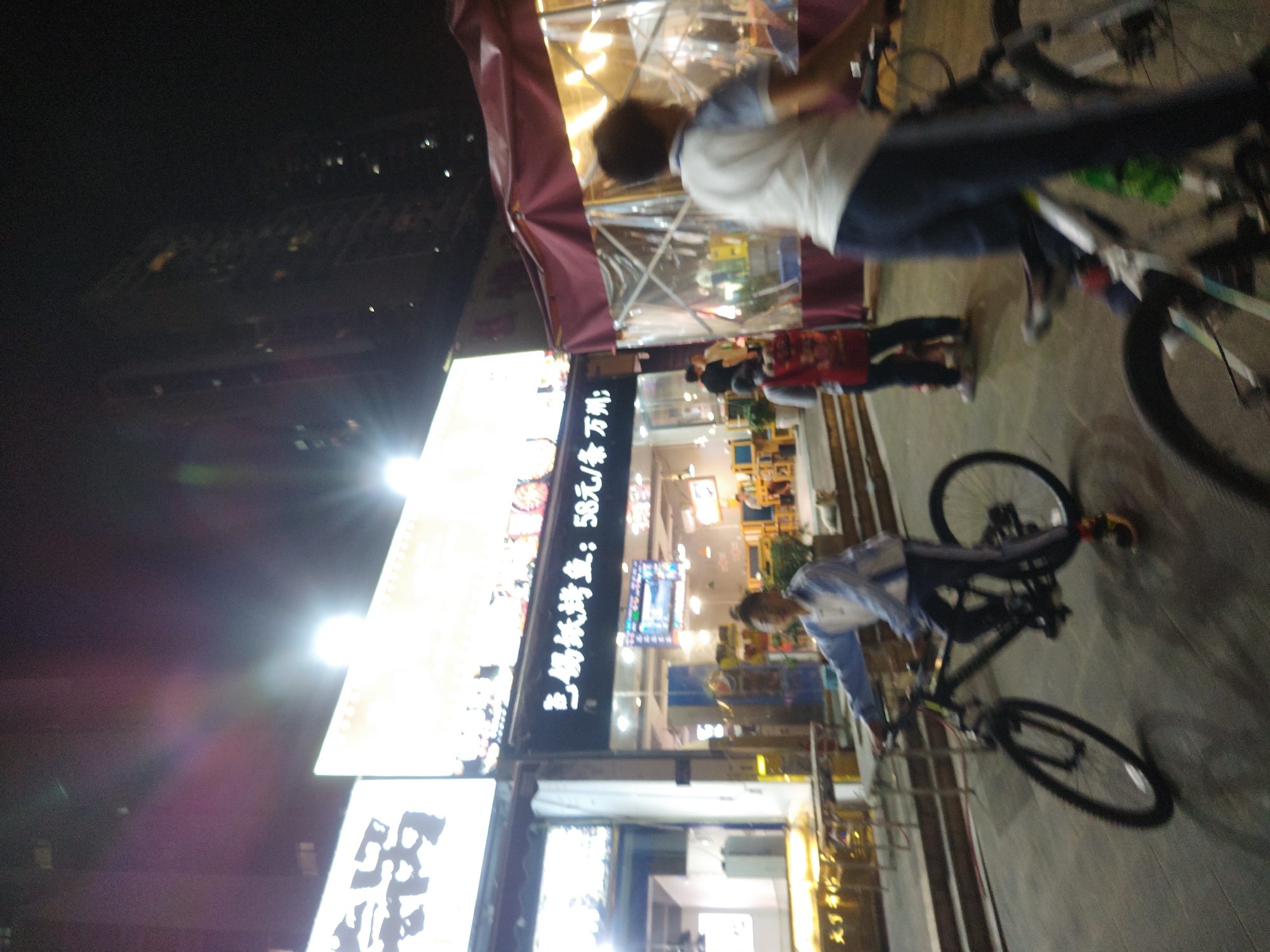 The image depicts a vibrant night scene on a commercial street in an urban area, likely Chongqing High-tech Zone. The sky is dark, illuminated by numerous bright lights from shops and streetlights, creating lens flares.
In the foreground, a person wearing a white and light blue long-sleeved shirt and dark pants stands beside a black mountain bike. Their body is partially turned, and they appear to be looking forward. Another bicycle is visible further to the right, suggesting a common mode of transport or activity in the area. Other blurred figures are seen in the background, including people inside a lighted establishment.
The left side of the frame is dominated by brightly lit vertical signs belonging to shops or restaurants. The most prominent sign displays Chinese characters, partially reading: "三码捞捞鱼:58元/斤 另万". This text likely refers to a dish or restaurant name, "San Ma Lao Lao Yu," with a price of "58 yuan per jin" (approximately 500g), followed by an incomplete phrase. Below this, another sign also features illuminated Chinese text, though it is less clear. Above these signs, a red awning extends over what seems to be an open-air or enclosed dining area, where multiple individuals are faintly visible through transparent barriers, indicating active business.
The scene suggests a lively evening, with people out on the street, and commercial establishments in full operation.