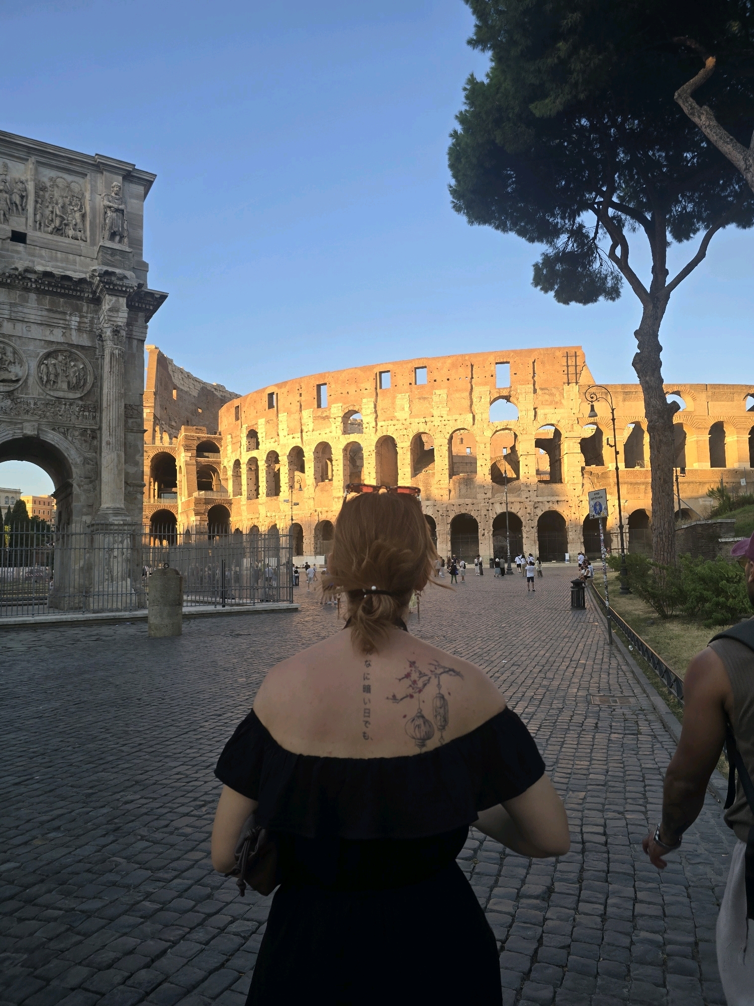 The image depicts a rear view of a woman walking on a cobblestone street in Rome, Italy, with the Arch of Constantine on the left and the Colosseum in the background. The woman has a tattoo on her upper back and is wearing a black off-the-shoulder dress. A man is visible to her right, also walking away from the camera. The sun is low in the sky, casting a warm glow on the ancient Roman structures. Several other people are walking in the distance, and trees and bushes are present along the path.