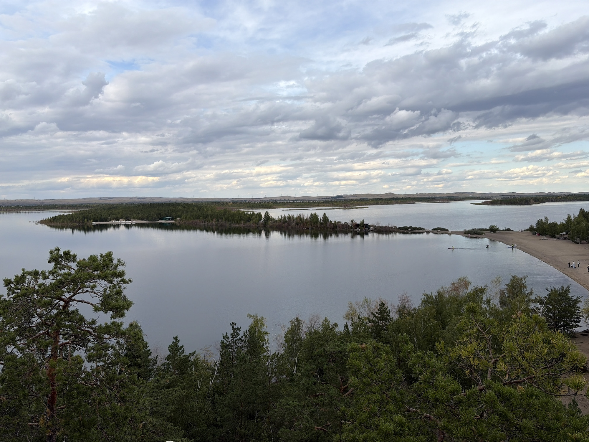 An elevated, wide-angle, medium shot captures a panoramic view of a large body of water, likely a lake, under a cloudy sky. In the foreground, dense green coniferous trees dominate the lower half of the frame, with a large pine tree on the left. The middle ground features the calm, grey water reflecting the sky and the distant treeline. On the right side of the water, a sandy beach is visible with a few people walking and two kayakers in the water. The background consists of a low, undulating treeline and hills stretching towards the horizon, under a sky filled with layered grey and white clouds, with patches of lighter sky breaking through. The overall impression is a natural, serene landscape.