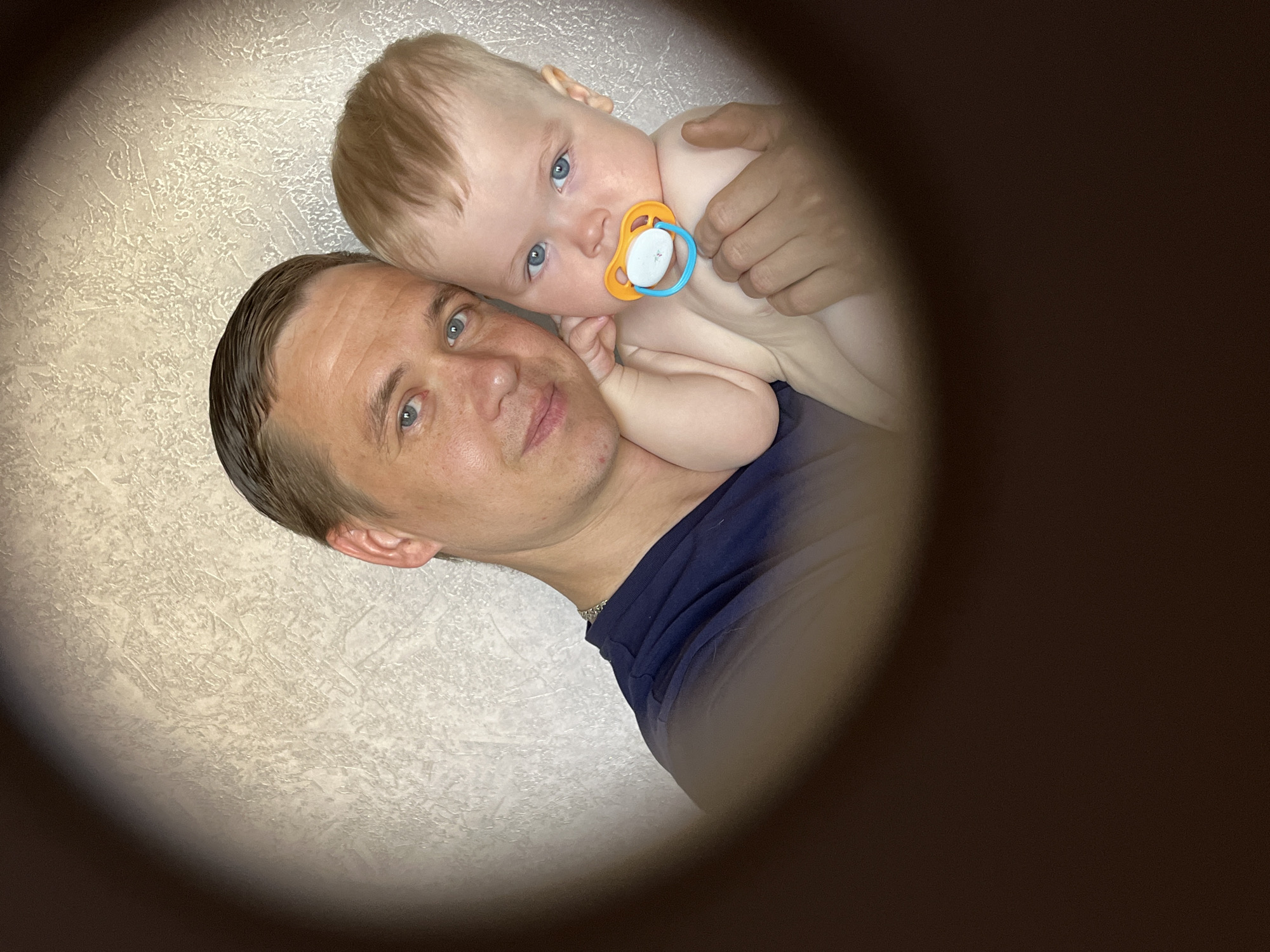 A close-up indoor photograph shows a man holding a baby. The man has short brown hair, fair skin, and blue eyes, and is wearing a dark blue shirt. The baby has blond hair, fair skin, and blue eyes, and is holding an orange and blue pacifier. The background is a white textured wall. The scene is viewed through a dark, circular border, giving a "keyhole" effect. There is no explicit indication of the location being in Chaikovsky, Russia.