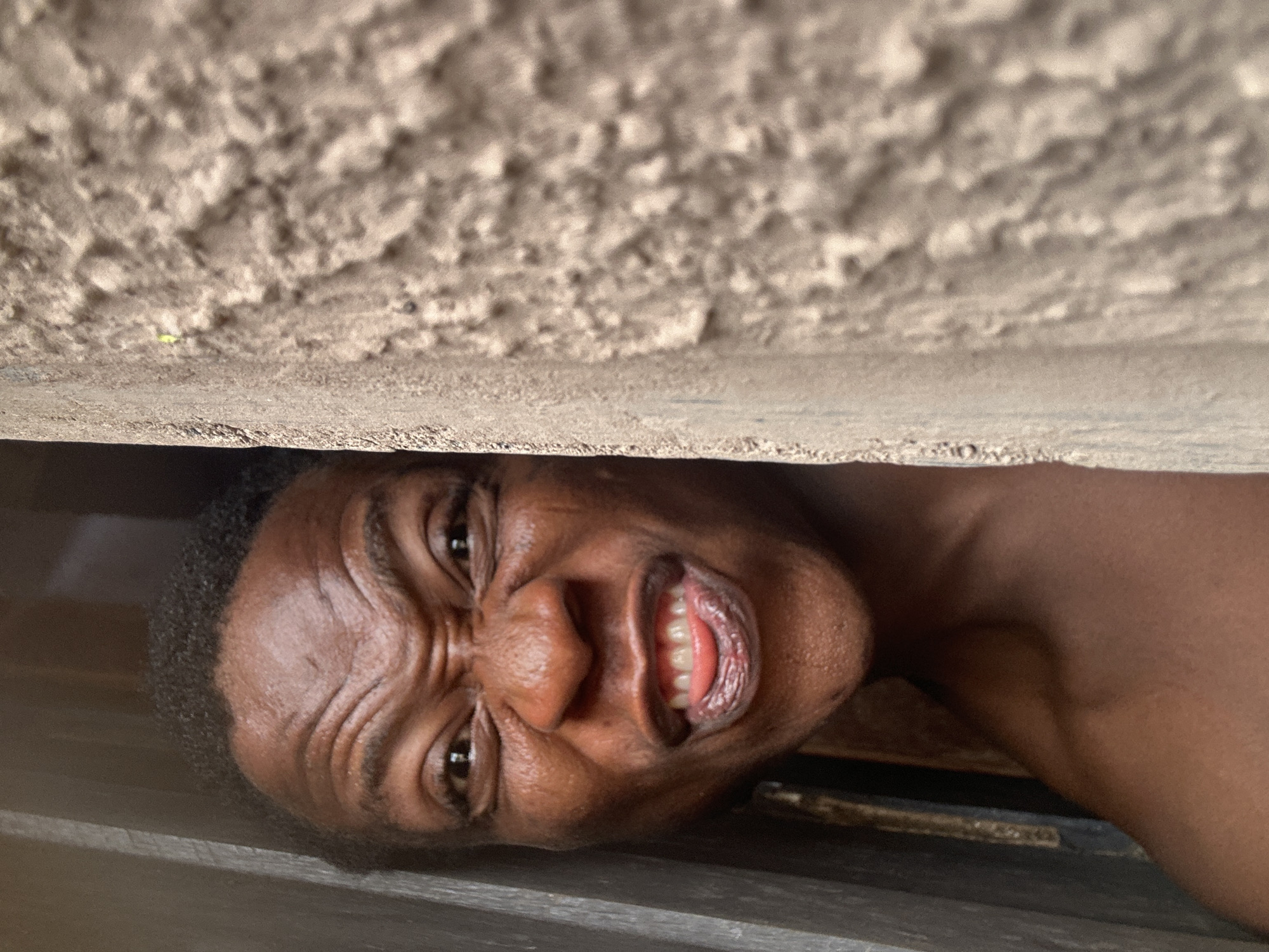 A close-up, slightly tilted view depicts a person with dark skin pressed between two surfaces. The upper surface is textured, possibly concrete or stucco, while the lower surface appears to be wood. The person's face is visible, with wrinkles and a strained expression. The background is out of focus. The location is not identifiable within Unknown, Unknown, based on the image.