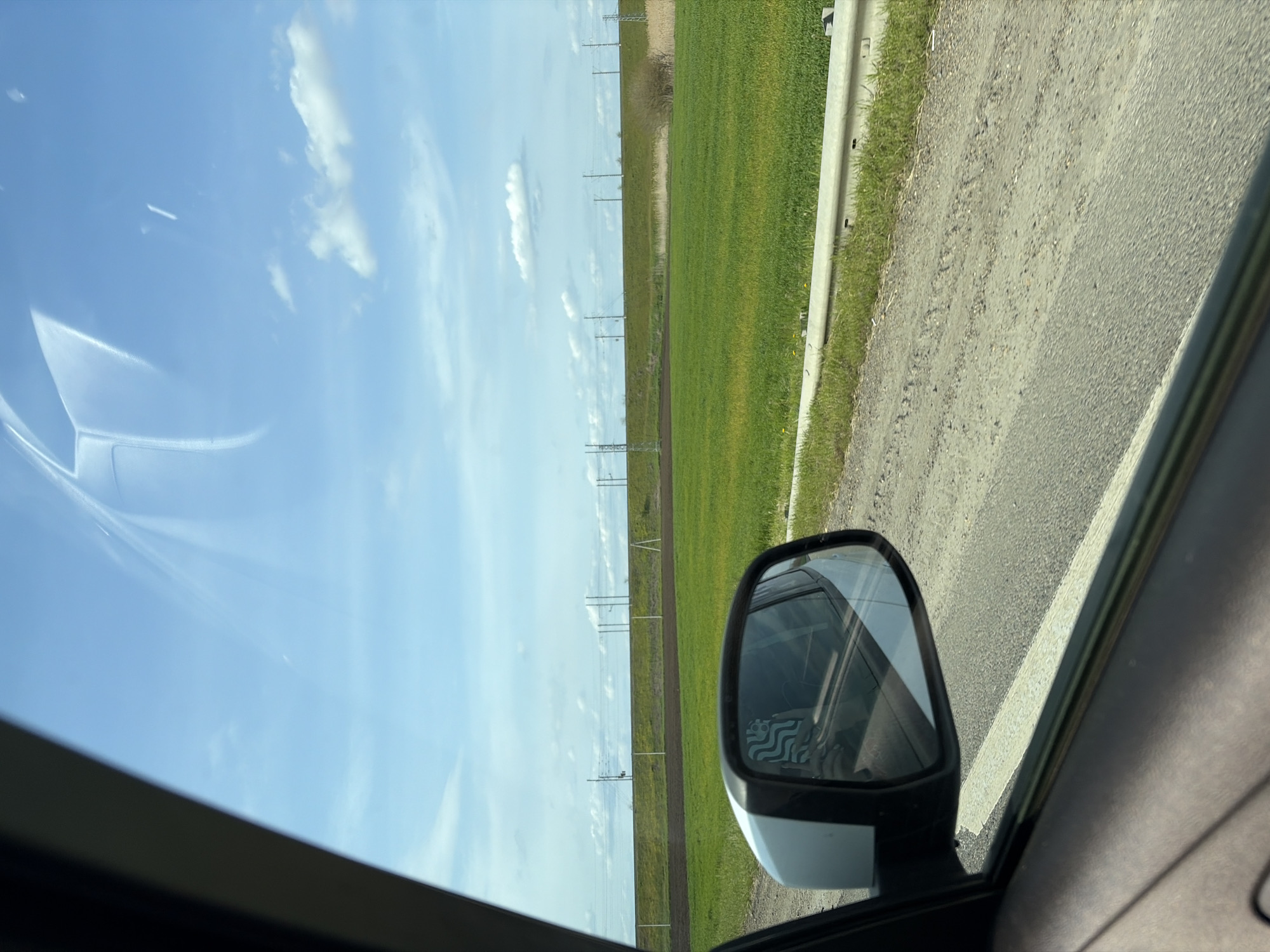 The image, correctly oriented horizontally, presents a view from inside a moving vehicle, looking out the passenger-side window across a rural landscape. The interior frame of the car window is visible along the left and bottom edges of the shot.

The upper portion of the scene is dominated by a clear, light blue sky with scattered white, fluffy clouds. Reflections of the sky, clouds, and some interior elements are visible on the car's window glass, particularly in the top left.

The lower part of the view shows a wide, green field extending towards the horizon under the bright sky. A series of utility poles or power lines stretches across the field into the distance, suggesting infrastructure in an open area. To the right of the field, a dirt or gravel road runs parallel to the car's direction, bordered by a concrete or metal guardrail with grass growing at its base.

A white side mirror of the car is prominently featured on the right side of the image, providing a reflection. Within the mirror's reflection, a blurred but identifiable stylized logo of a siren or mermaid, resembling the Starbucks emblem, can be discerned, likely from an object inside the car.

The scene depicts a bright, sunny daytime, with clear weather conditions, and appears to be in a rural or semi-rural setting. The green fields suggest a temperate climate, possibly late spring or summer. The context of Medvyodovskaya, Russia, aligns with the open, agricultural landscape. There is no other legible text visible besides the faint logo in the mirror.