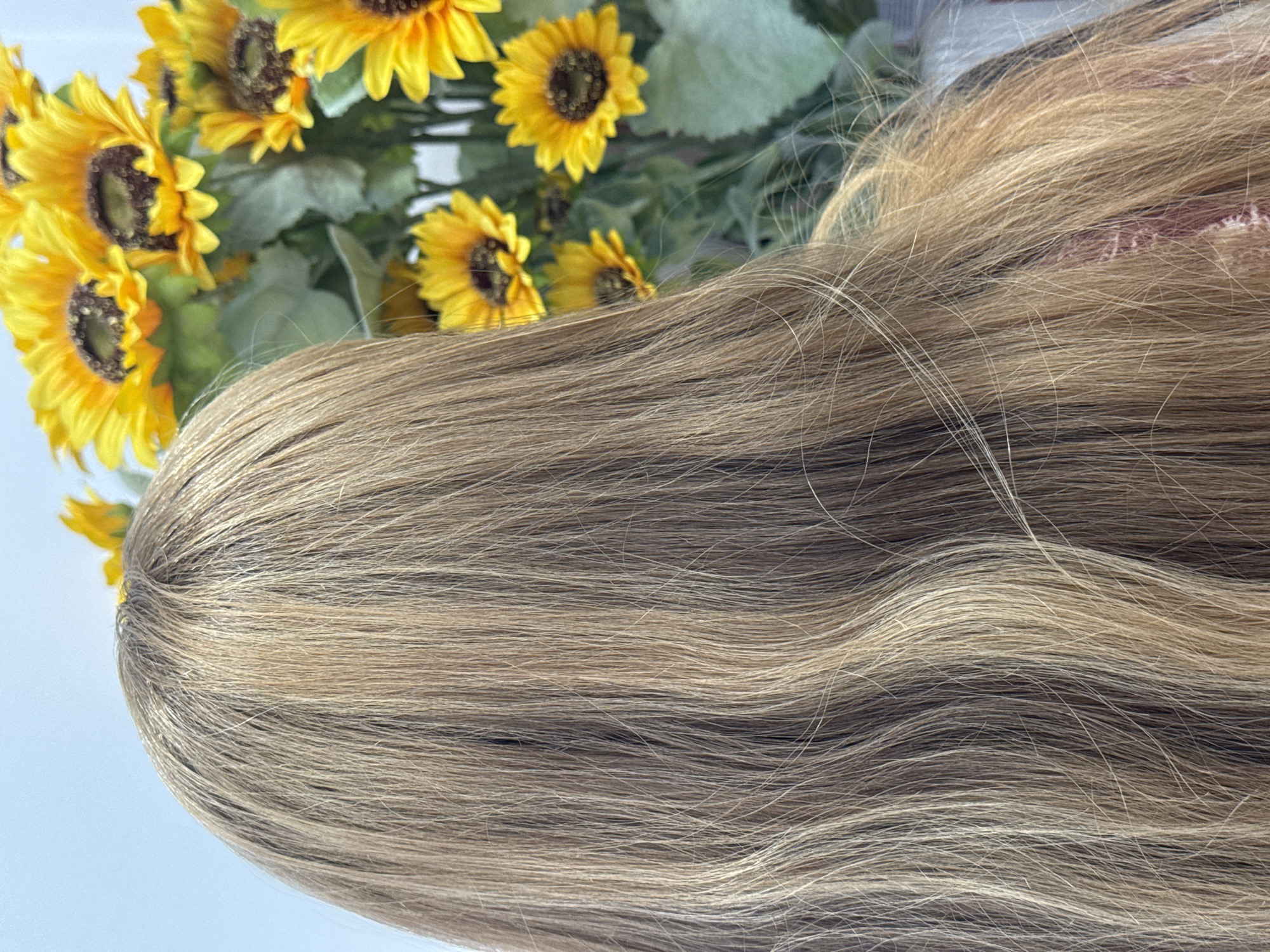 The image shows the back of a person's head with their long, blonde, wavy hair. In the background, there is a bouquet of bright yellow sunflowers and green leaves, placed against a white wall. The scene is likely indoors, given the lighting and background elements. There is no discernible location in the city of Podgorica, Montenegro.