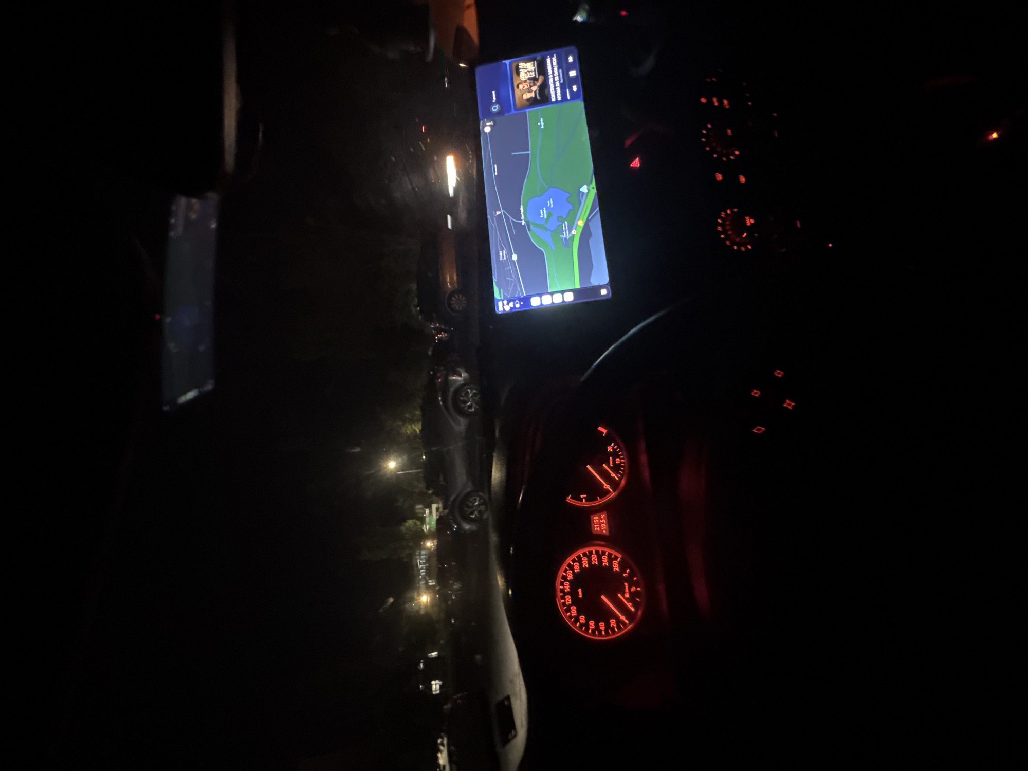 The image shows the interior of a car at night, with a mobile phone mounted on the dashboard displaying a map showing a location in Stara Zagora, Bulgaria. The map includes an area of green space with a body of water (likely a lake or pond). The car's dashboard is illuminated, showing the speedometer and other gauges. The speedometer reads 0 km/h. The outside setting through the windshield shows a street lit by streetlights with some cars parked to the side. The time shown on the dashboard is 21:56, with an indicated temperature of +19.5°C.