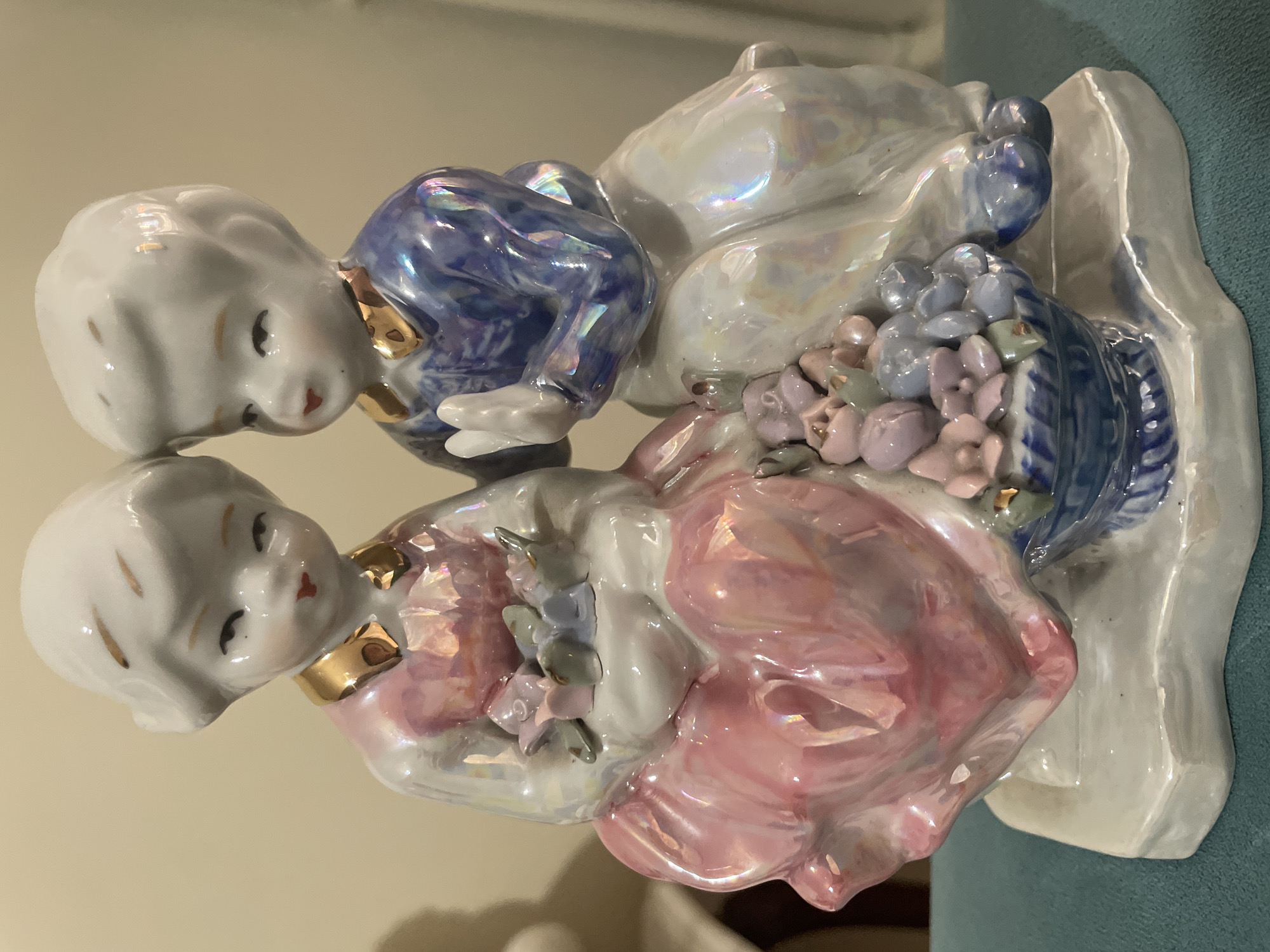 This is a close-up, eye-level studio photograph of a porcelain figurine depicting a boy and a girl sitting side-by-side on a white base. The figurine has an iridescent glaze, giving it a pearlescent sheen that reflects light in various colors. The girl, on the left, wears a pink dress with gold trim around the collar and is holding a small bouquet of flowers. Her hair is styled in a bob. The boy, on the right, wears a blue outfit with a gold collar and is holding his hands clasped in front of him. He also has a gold trim on his collar. Between the two figures, at their feet, is a basket filled with colorful flowers in shades of pink, blue, and purple. The background is a plain, light-colored wall. The figurine is placed on a teal surface. There is no discernible connection to Kadıköy, Turkey, in the provided image.