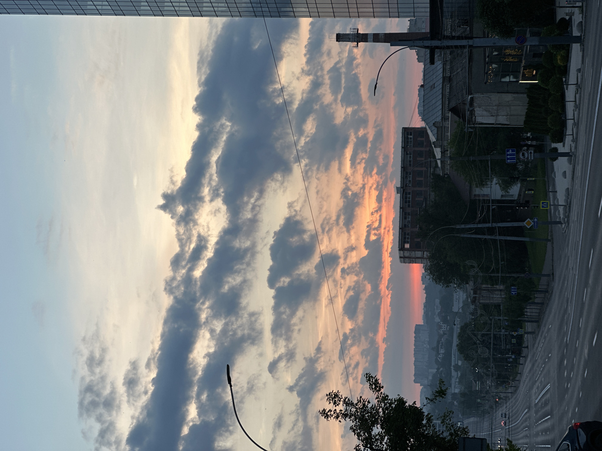 This image presents an elevated urban landscape in Chișinău, Moldova, captured during either sunrise or sunset, with a general southeast orientation. The sky occupies the upper two-thirds of the frame, displaying a gradient from light blue at the top, transitioning to pale yellow and then to a vivid orange-red glow concentrated at the horizon. This illumination is partially filtered by dense, dark gray stratocumulus clouds that are arranged in undulating layers across the mid-sky.
In the foreground and mid-ground, a multi-lane asphalt road, likely Strada Alexandru cel Bun, extends from the lower left, gently curving and descending towards the center-right. It features clear white lane markings, including double solid lines and dashed lines, and appears relatively devoid of traffic, with only a few distant vehicles visible. A dark-colored vehicle is partially visible in the bottom left corner. Along the left side of the road, a tall, curved street lamp pole extends into the upper-left sky. Below it, the upper foliage of a tree is silhouetted against the clouds. Various traffic signs are visible along the road, including blue directional signs, a pedestrian crossing sign, a yellow diamond warning sign, and a blue 'no trucks' sign. Decorative metal scrollwork is attached to several street light poles along the road.
On the right side of the image, a substantial section of a modern, tall glass skyscraper is visible, identifiable as the Kentford Business Centre (located at Blvd. Ştefan cel Mare şi Sfînt 202). Its reflective facade displays a grid pattern. To its left and slightly foreground, a tall, slender industrial smokestack or chimney, featuring a red and white pattern, rises prominently. Below these larger structures, a collection of diverse buildings populates the street level; these include older, multi-story European-style structures, some appearing residential, and one building in the mid-distance showing exposed framework or scaffolding on its upper floors. A building in the lower right corner features a distinct, vertical, illuminated sign.
The distant background reveals a hazy urban skyline composed of numerous residential buildings, silhouetted against the intense red-orange horizon. Sparse trees and shrubs are interspersed throughout the urban environment. The scene is devoid of observable individuals or direct human interaction, conveying a quiet, transitional moment within the city.