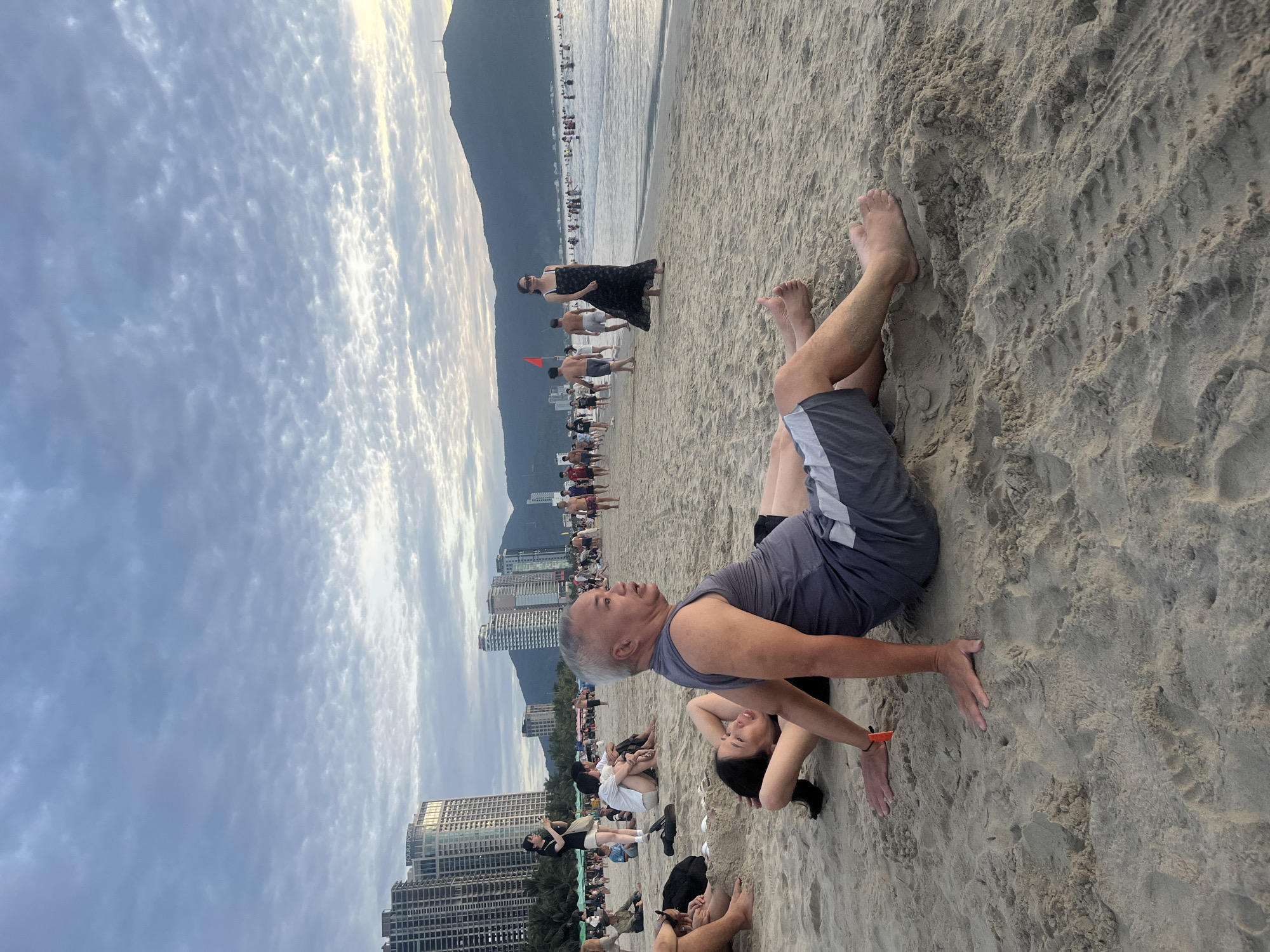 The image captures a bustling beach scene at My Khe Beach in Đà Nẵng, Vietnam, identifiable by the presence of the "Four Points by Sheraton Đà Nẵng" hotel among other high-rise buildings in the background and the distinctive Sơn Trà Peninsula mountains to the right. In the foreground, an East Asian man with short grey hair, dressed in a grey sleeveless shirt and shorts, sits on the sand, facing towards the right side of the frame with a slight upward gaze. Behind him, an East Asian woman with dark hair lies supine on the sand, her arms bent with hands behind her head, looking towards the man. The sandy beach is populated by numerous individuals; to the mid-left, a young East Asian woman stands holding a smartphone, seemingly capturing an image, while other groups sit or relax on the sand. To the mid-right, an East Asian woman in a long black dress and sunglasses walks along the shoreline, facing towards the viewer. The calm ocean in the background contains numerous people swimming and wading, with a red flag visible in the water, indicating a designated area. The horizon is dominated by modern high-rise buildings and a prominent dark mountain range featuring two small antennae-like structures on its peak. The sky is overcast with scattered, textured clouds, allowing some light to penetrate.