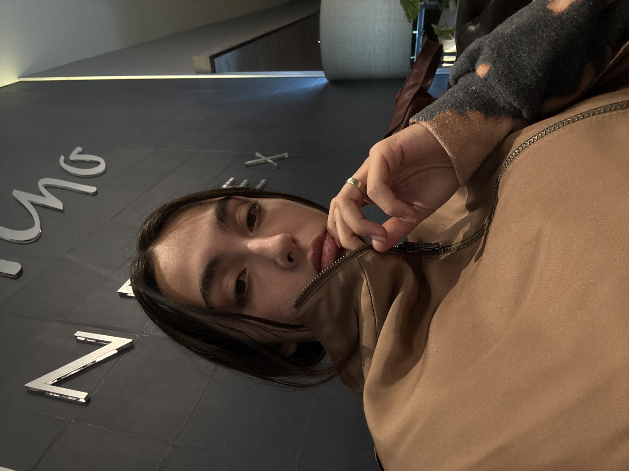 A person is positioned in front of a dark-gray wall that has what looks like polished silver letters fixed to it, perhaps a sign. The person's upper body is covered in a brown-colored zipper jacket and a gray long-sleeved shirt that has some discoloration around the wrist. On their left hand is a simple gold ring. They are looking at the camera with a slight smirk. Behind them is a dimly-lit space, with a large concrete or stone cylinder. While the text is not clear from the image alone, it is possible that the sign on the wall reads "Auditorio Nacional", which is a cultural venue in Mexico City, Mexico.