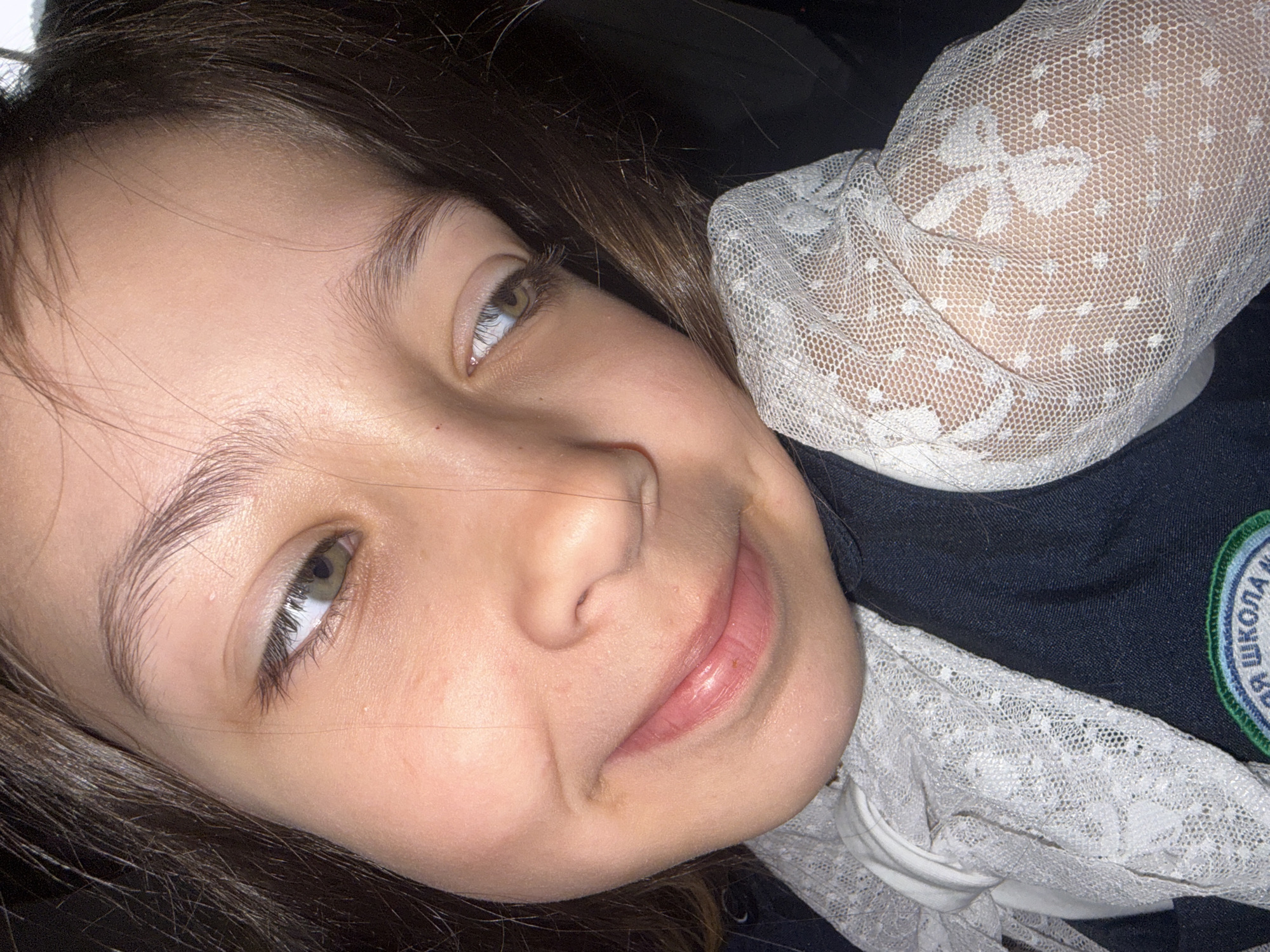 A close-up, angled headshot depicts a young girl with light brown hair and fair skin. Her eyes are open, gazing slightly upward, and she exhibits a faint smile. She is wearing a school uniform consisting of a dark blue jacket and a white lace collar and sleeves. The uniform bears a patch with text in Russian, indicating it is from a school. Based on the text, the school is in Votkinsk, Russia. The background is dark and indistinct.