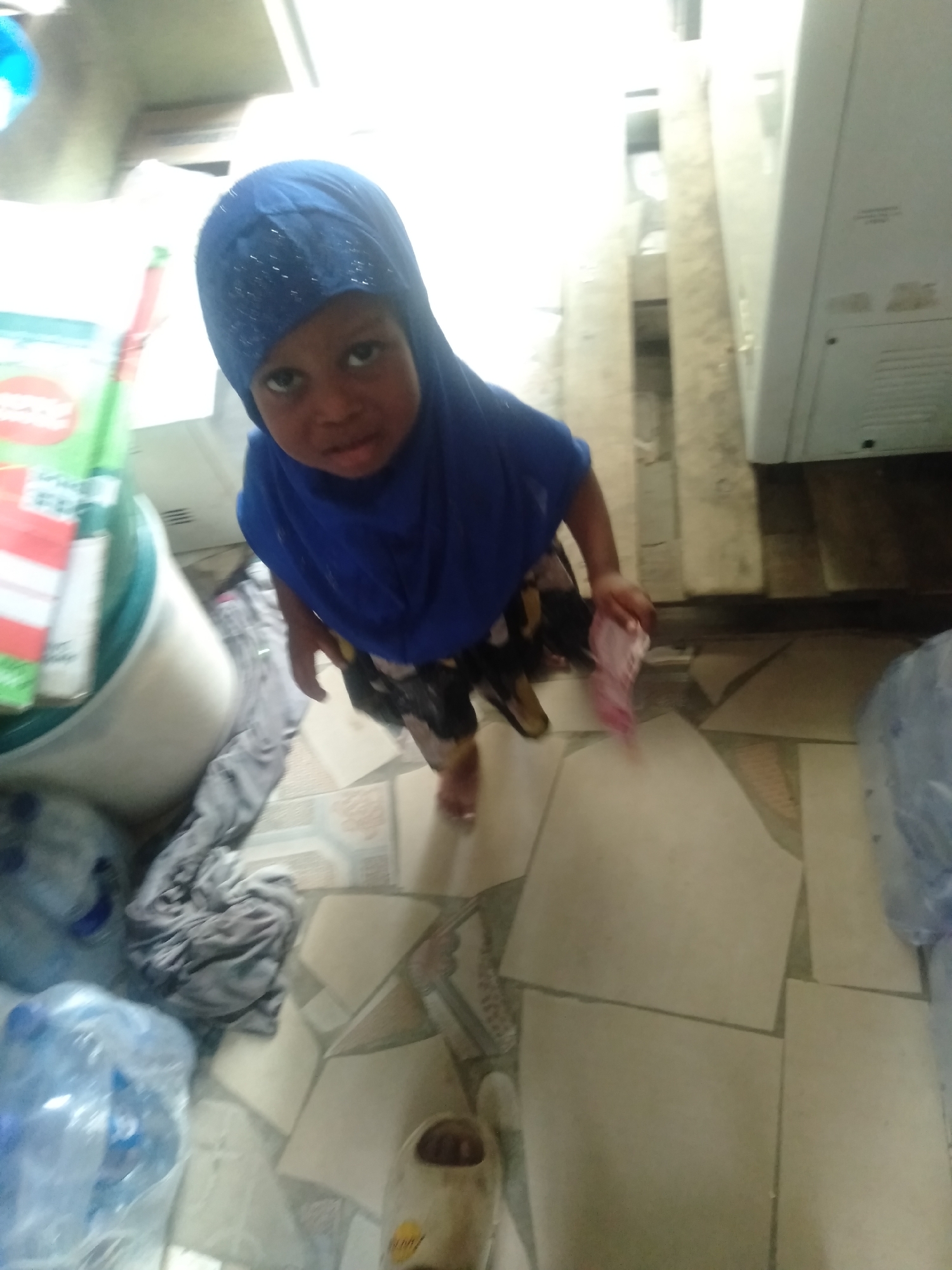 A high-angle, slightly blurry image, taken in Shangisha, Nigeria, captures a young child looking directly at the camera. The child is wearing a blue head covering (hijab), a blue top, and patterned shorts. They appear to be holding a small, light pink item in their right hand.
The setting is an indoor space, possibly a utility room or storage area, with a floor covered in irregularly laid beige and patterned tiles. To the left of the child, there's a white bucket or container, a folded gray textile (possibly clothing or a blanket), and multiple large plastic bottles, some filled with water. A green and red box is partially visible near the bucket, but any text on it is illegible. In the background to the right, a white household appliance, resembling a washing machine or refrigerator, stands against a wall. A brown/beige slipper or shoe is partially visible on the floor in the foreground. The lighting is consistent with an indoor environment. The child's facial expression is neutral.