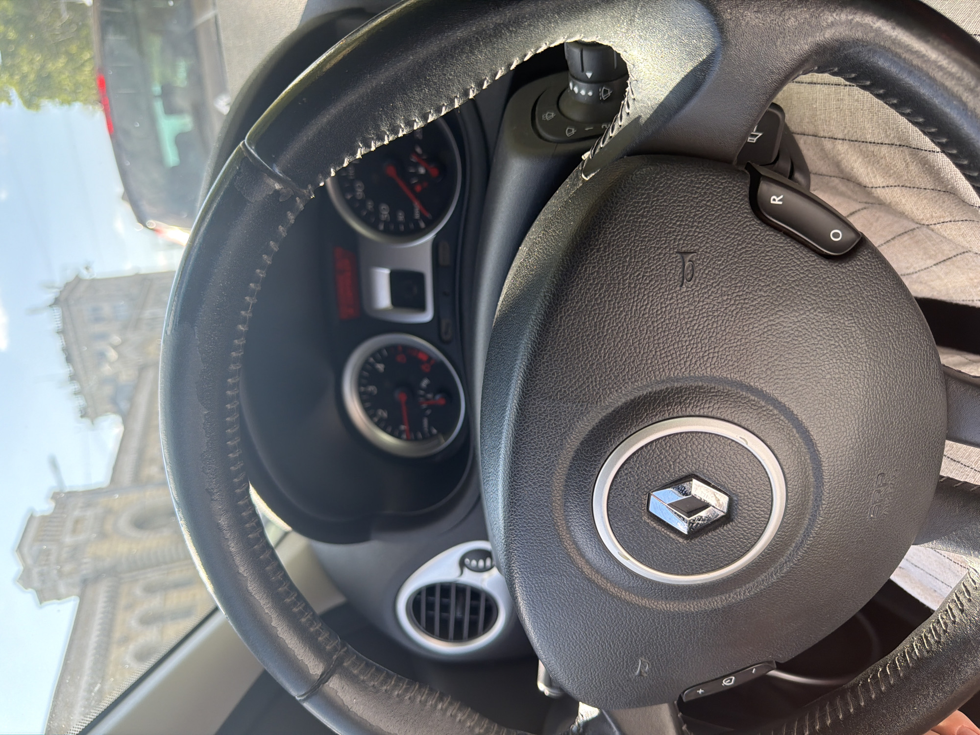 The image shows the interior of a car, with a focus on the steering wheel and dashboard. The steering wheel is black leather with visible wear and stitching. The Renault logo is prominently displayed in the center of the steering wheel. To the left of the steering wheel, there is a horn button, and to the right, there are controls for the windshield wipers and turn signals. The dashboard includes two circular gauges with red needles, likely for speed and RPM, and a digital display. A circular air vent with a silver trim is visible to the left of the gauges. Outside the car, through the windshield, a building with neoclassical architecture and a clear blue sky are visible. A dark-colored vehicle is also seen in the background. The overall impression is that the car is stationary, likely parked or stopped in traffic. The light suggests it is daytime. The background architecture appears to be from Chișinău, Moldova, possibly near a historical district.